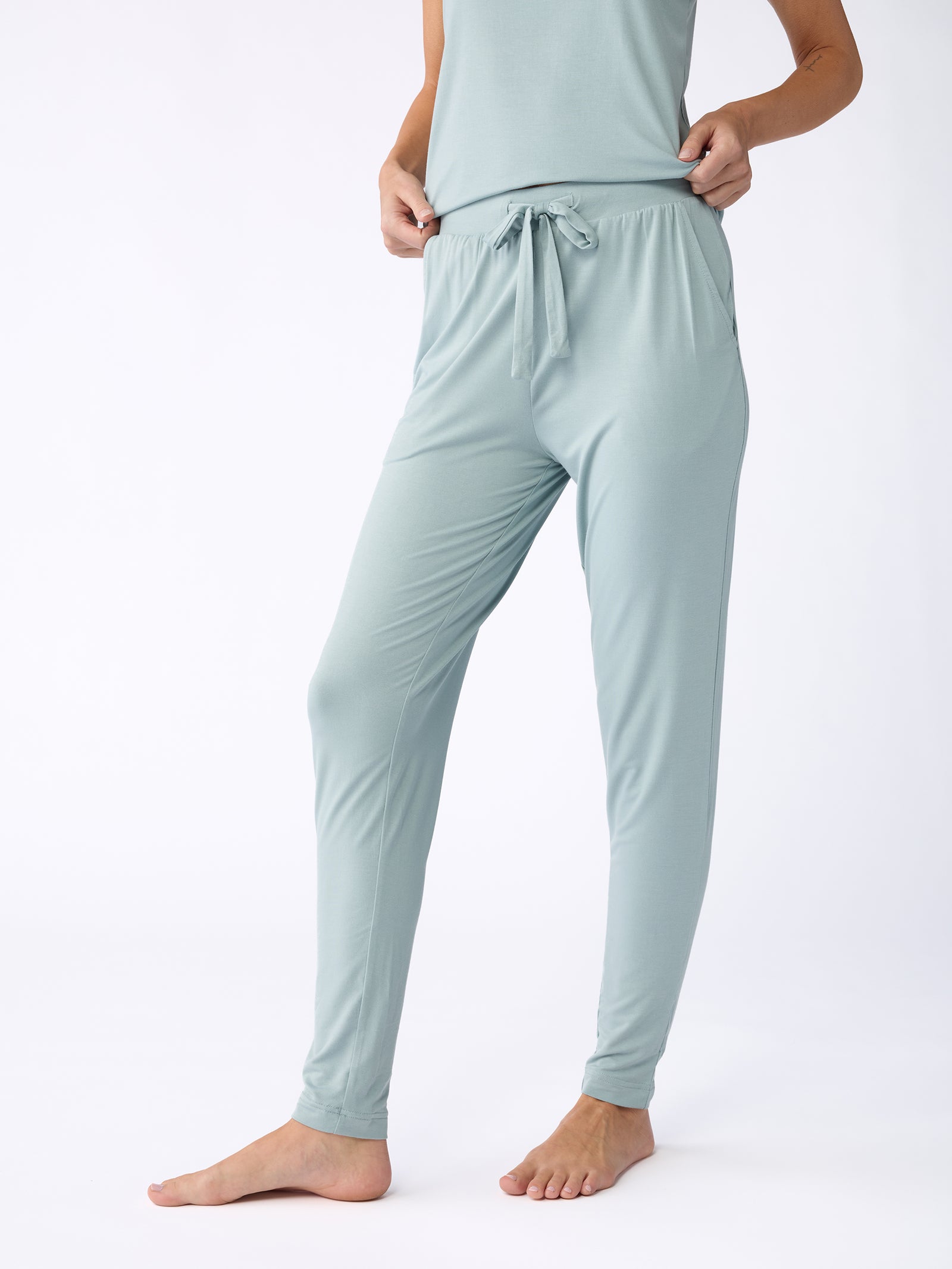 A person wearing Cozy Earth's Stretch Knit Bamboo Tapered Pant in light blue stands barefoot against a plain white background, with the image focusing on the lower body to highlight the pant's fit and design. 