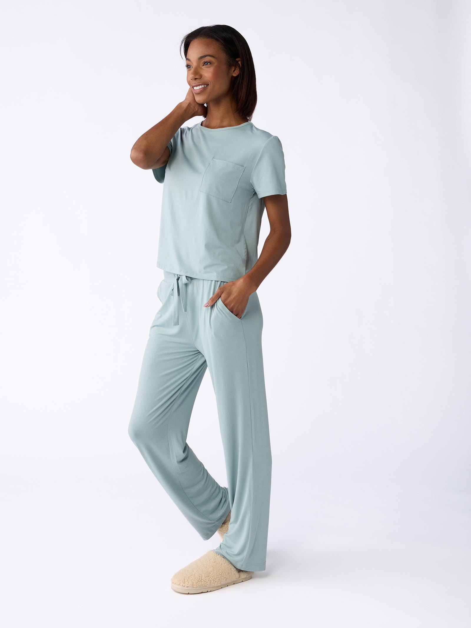 A person smiles while wearing the Women's Stretch-Knit Bamboo Lounge Tee from Cozy Earth, which is a light blue loungewear set with a short-sleeve top and long pants. They have their hand resting on their hip and are standing against a plain white background, wearing beige slippers. 