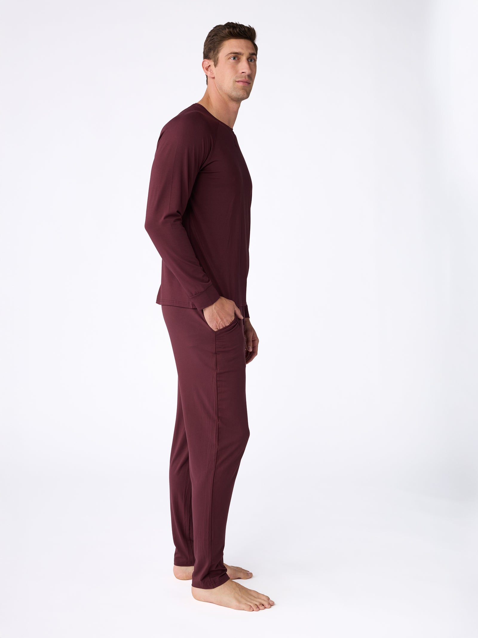 A man wearing the Cozy Earth Men's Stretch-Knit Bamboo Long Sleeve pajama set in a burgundy hue stands barefoot against a plain white background, gazing upward and to the left with his hands tucked into his pockets. 