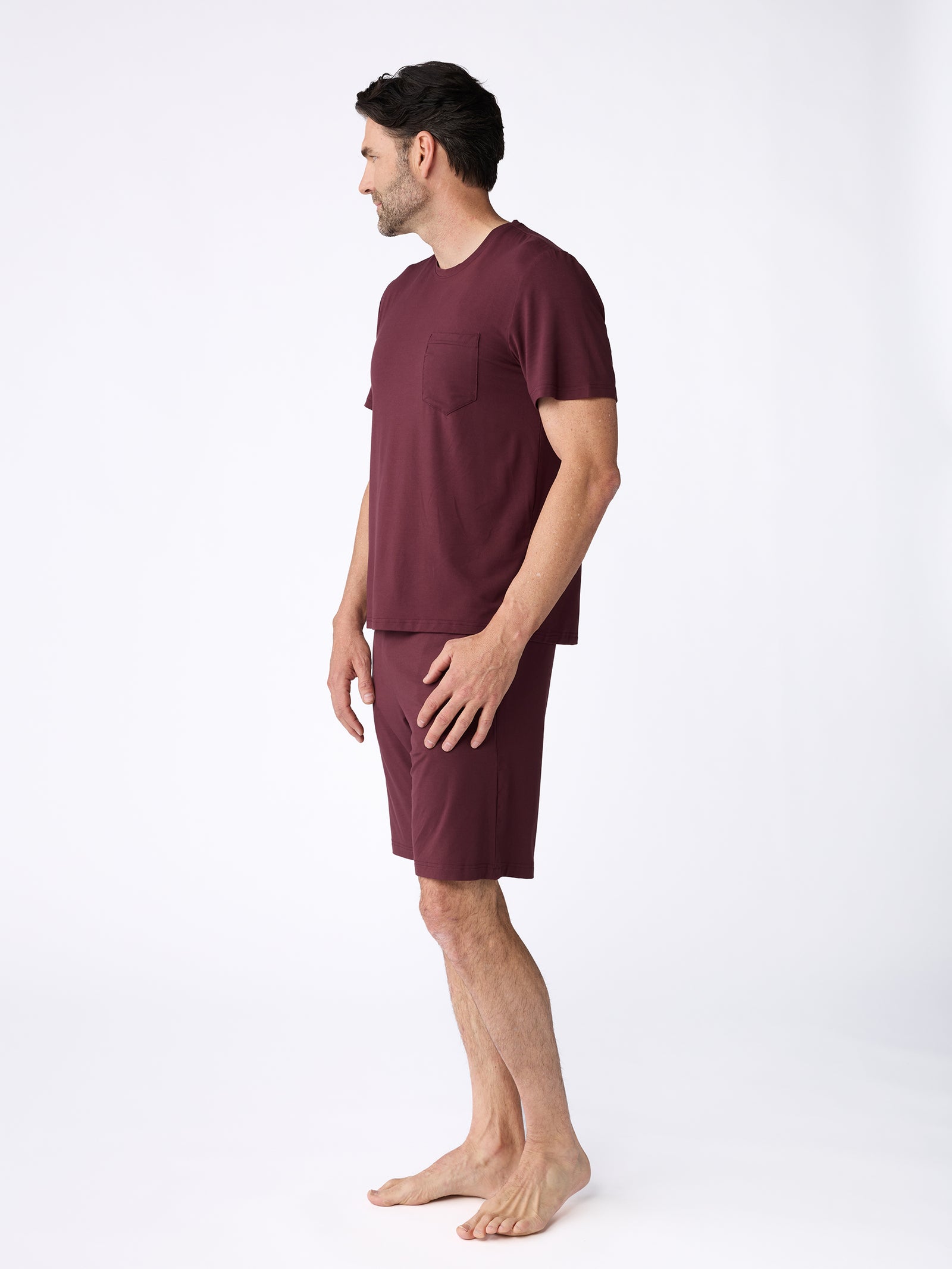 A man stands barefoot against a plain white background, wearing a matching set of Cozy Earth's Men's Stretch-Knit Bamboo Pajama Short and a maroon t-shirt. He is looking to his left with a relaxed posture. 