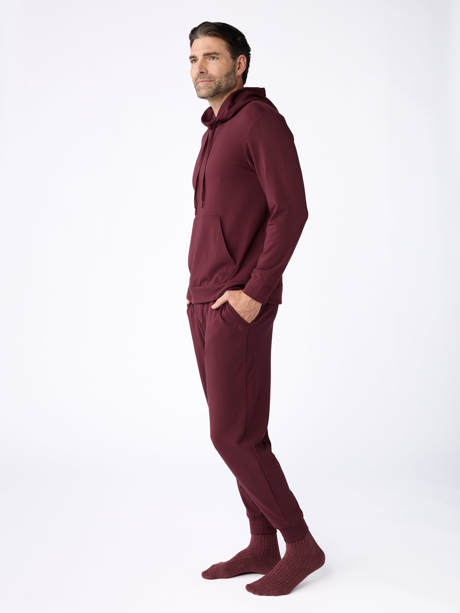 A man dressed in Cozy Earth's Men's Ultra-Soft Bamboo Hoodie, paired with matching pants and socks in rich dark red, stands against a plain white background. He gazes to his left with his hands casually tucked into his pockets, exuding a relaxed posture. 