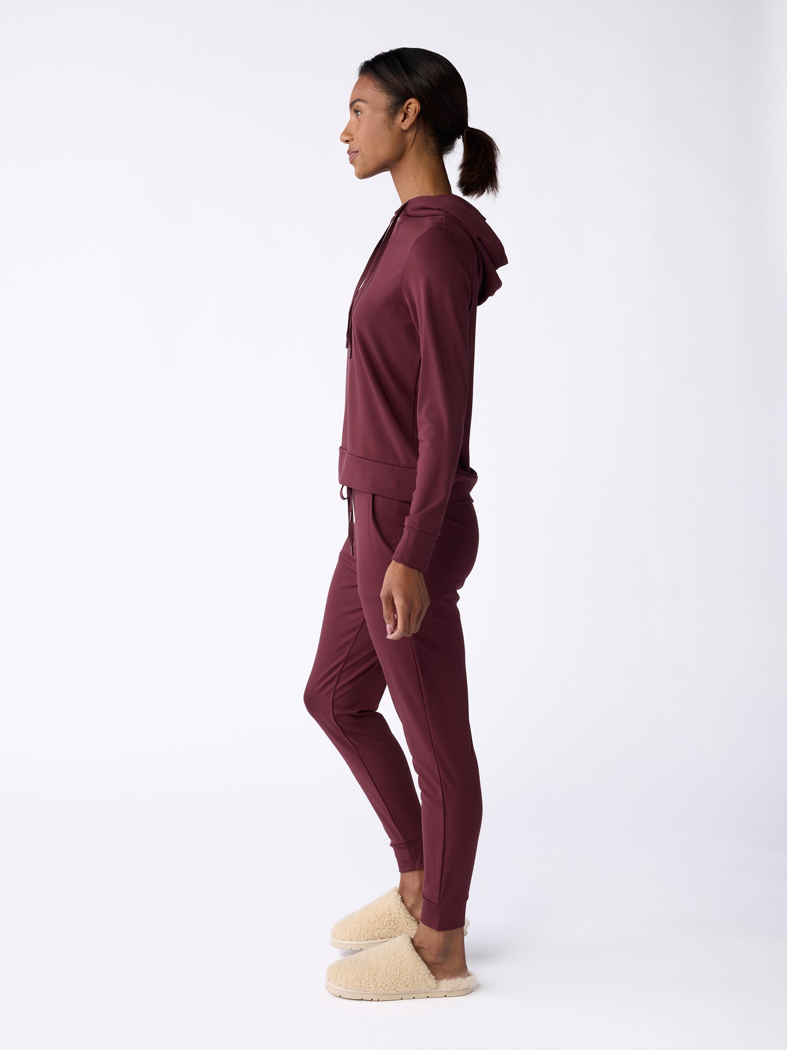 A person is standing in profile against a plain backdrop, wearing a maroon Women's Ultra-Soft Bamboo Hoodie by Cozy Earth, paired with matching jogger pants and beige slippers. The image highlights the side view of the outfit. 