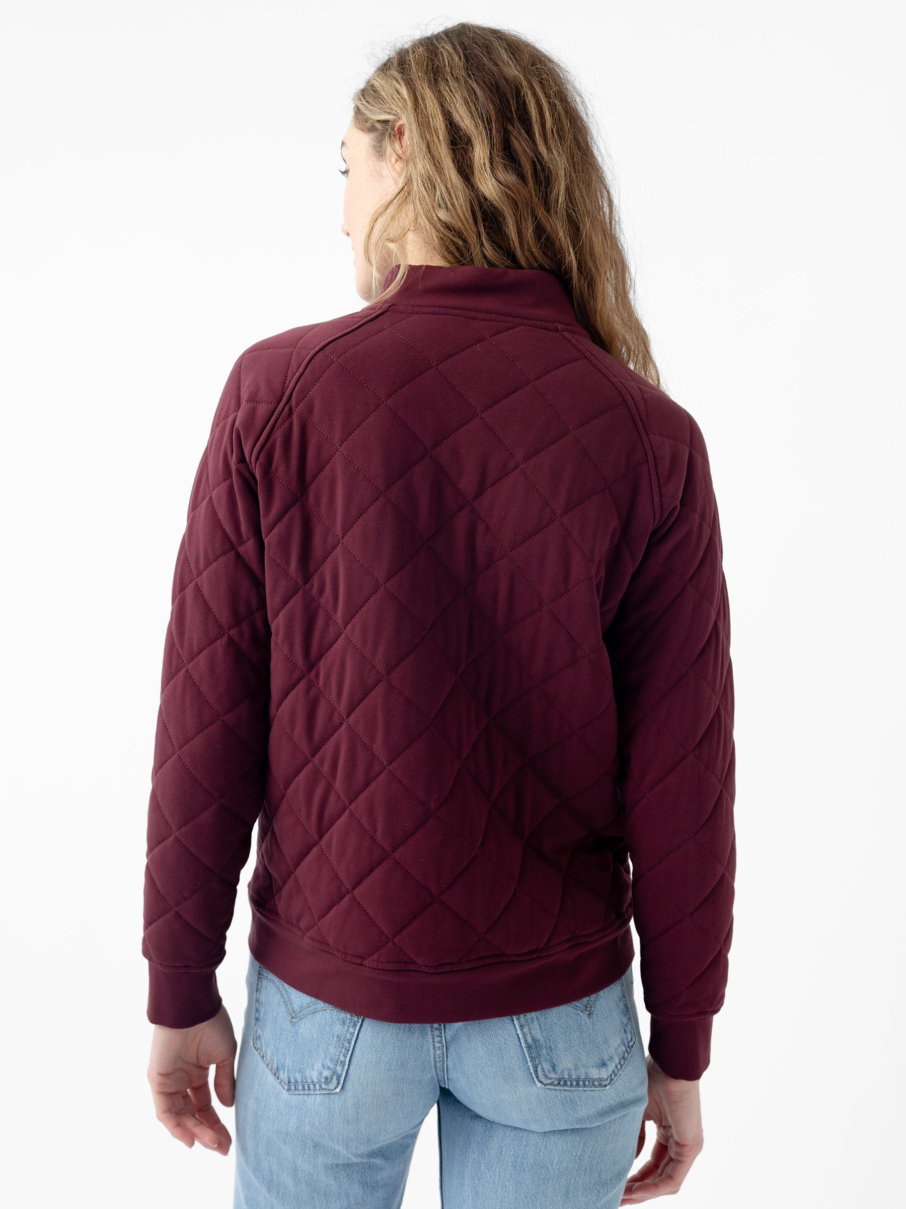 A person with long, wavy hair is standing with their back to the camera, wearing a maroon Women's Ultra-Soft Bamboo Quilted Snap Pullover by Cozy Earth over light blue jeans. The background is plain white. 