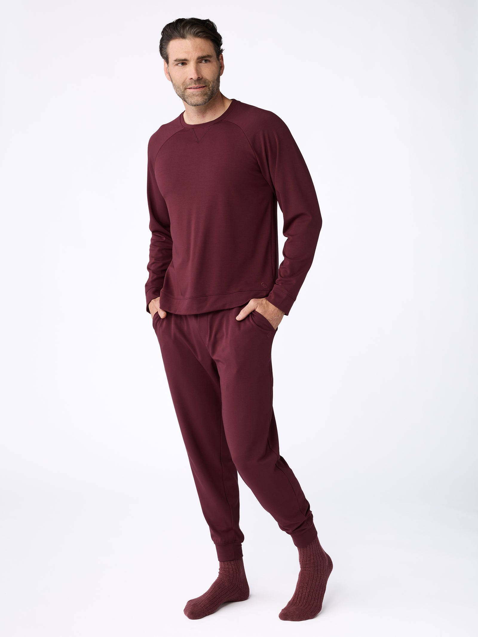 A person is standing in a relaxed pose, wearing the Men's Ultra-Soft Bamboo Pullover Crew from Cozy Earth in burgundy, paired with matching pants and knitted socks. The background is plain white. 