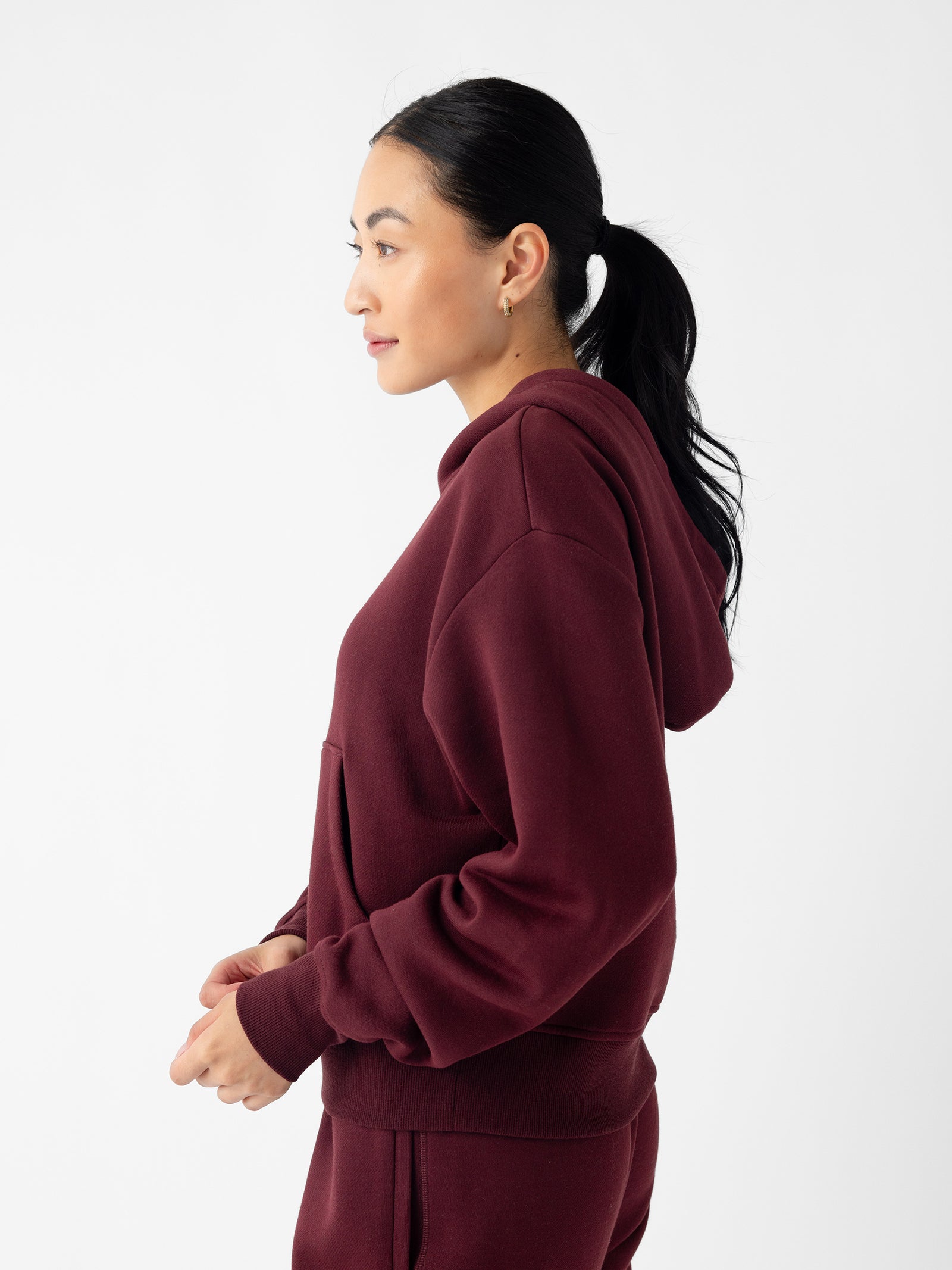 A person with long dark hair tied in a ponytail is seen in a profile view, wearing the Cozy Earth Women's CityScape Cropped Hoodie in maroon and matching pants. The background is plain white, and the person is looking to the left with a relaxed expression. 