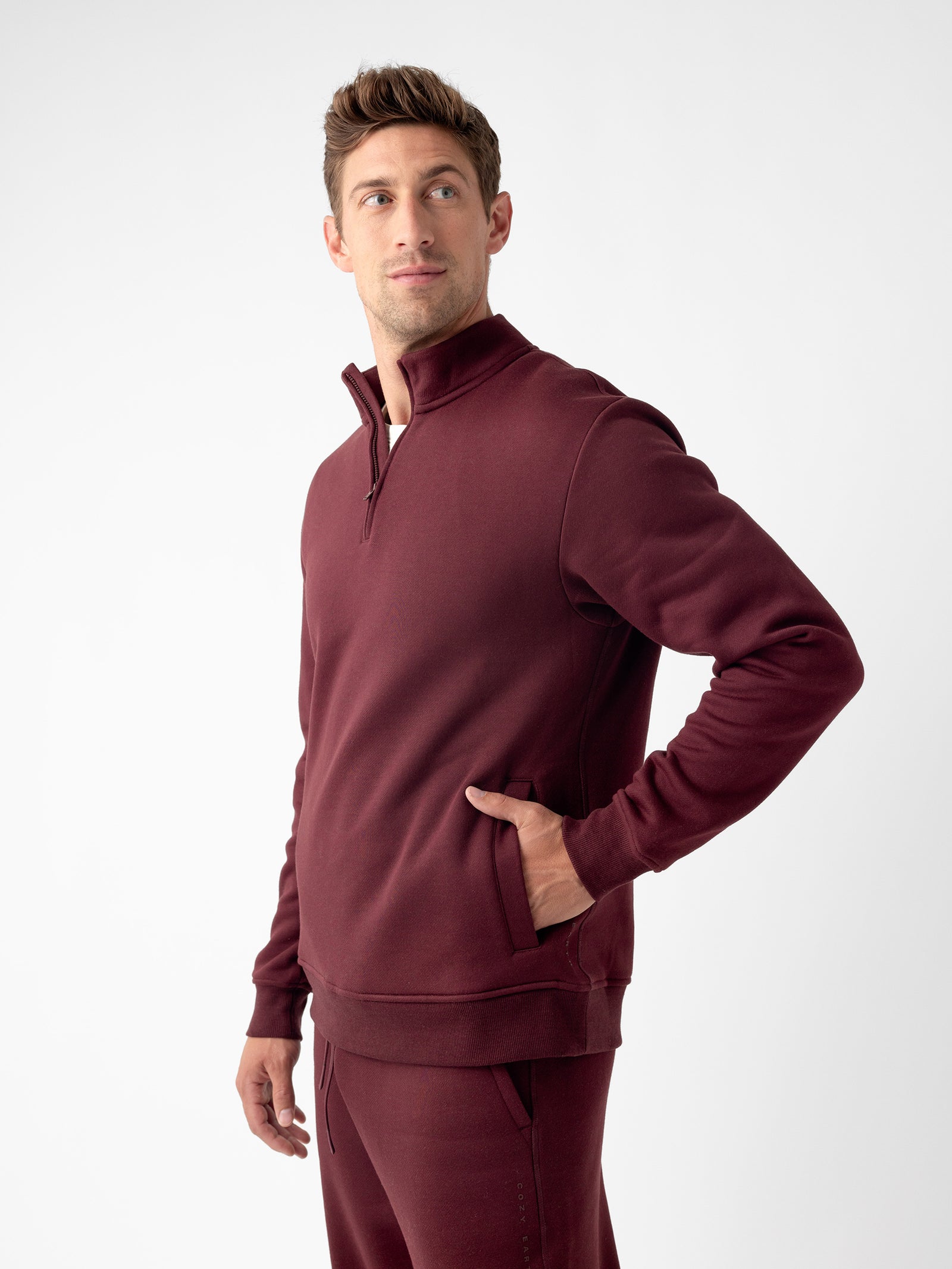 A person wearing a matching maroon Cozy Earth Men's CityScape Quarter Zip and pants is looking upwards and to the side. The individual has their right hand in the pocket of the sweatshirt. The background is plain and white. 