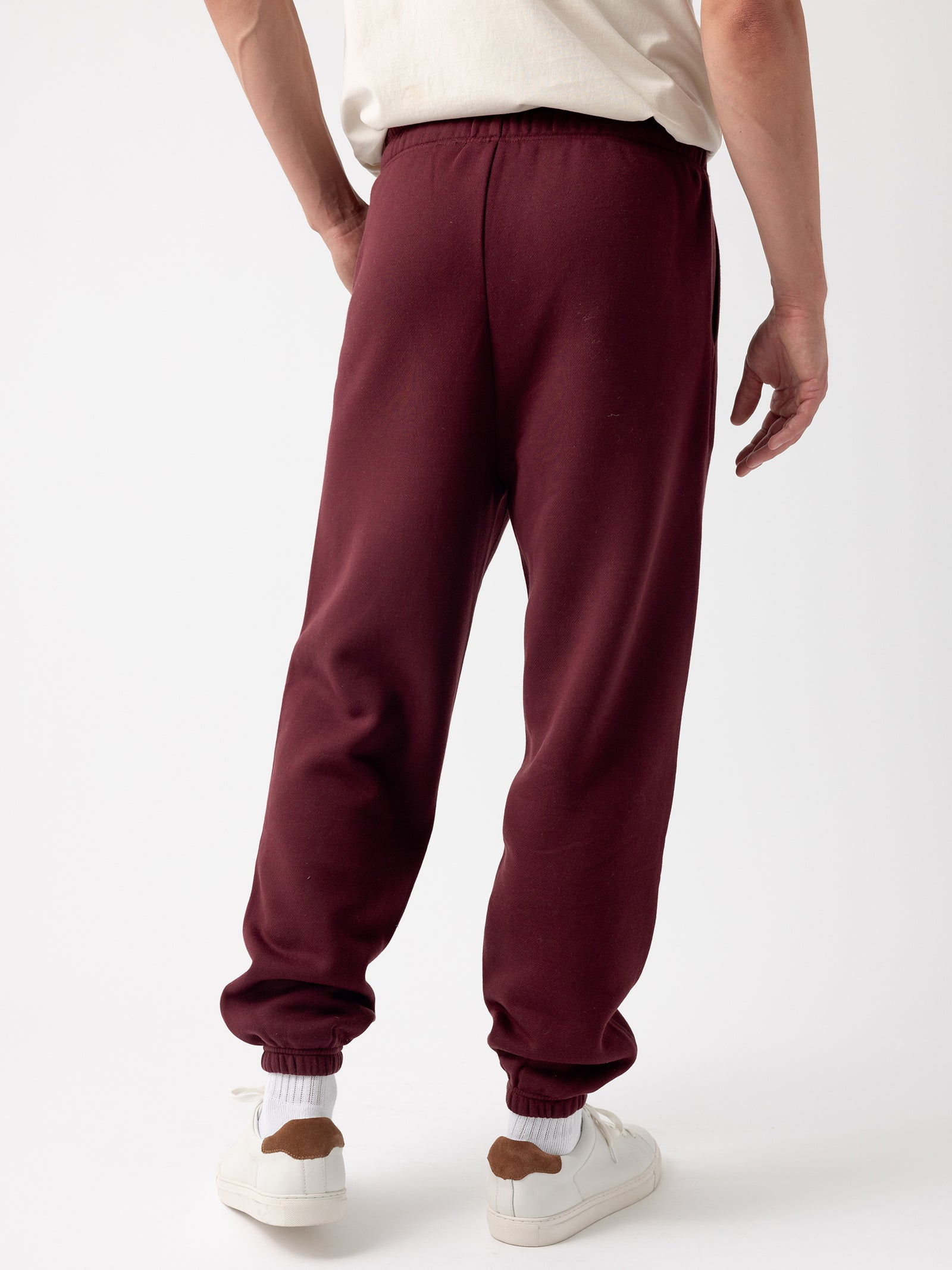 A person in a white shirt stands with their back to the camera, wearing the Cozy Earth Men's CityScape Sweatpant in maroon, which features elastic cuffs, paired with white sneakers that have brown accents. The background is plain white. 