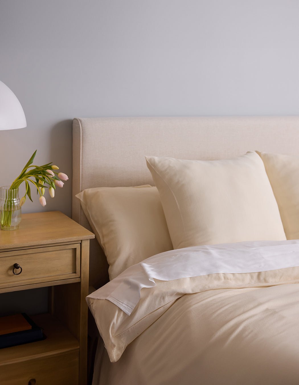 A neatly made bed with Cozy Earth's Aire Bamboo Shams in cream and pillows sits beside a wooden nightstand featuring a small lamp and a clear vase of pale pink tulips, set against a light gray wall.