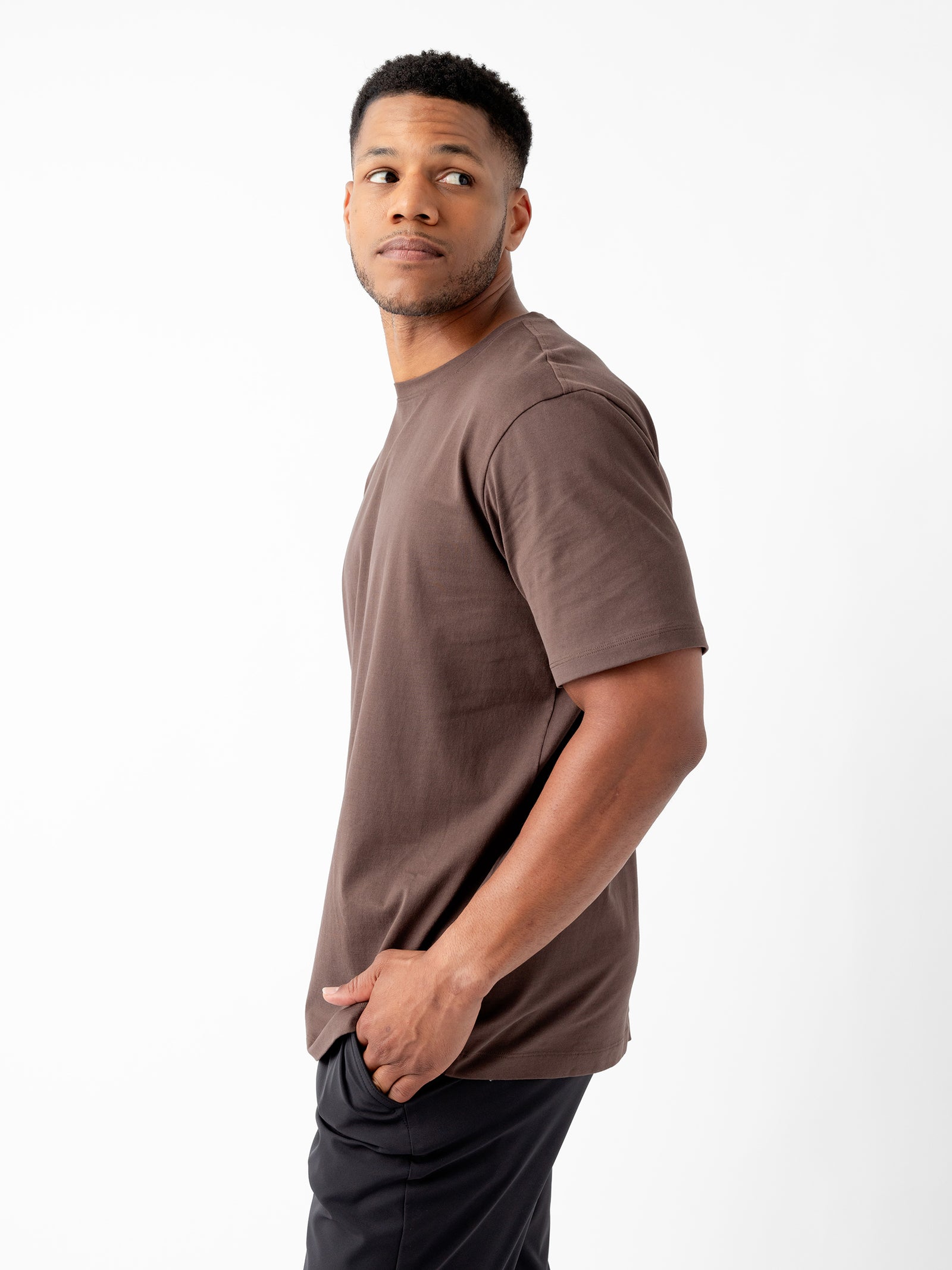 A person with short curly hair and a beard is seen in a side profile, looking slightly over their shoulder. They are wearing the Men's All Day Tee by Cozy Earth, a brown short-sleeve T-shirt, along with black pants and one hand in their pocket. The background is plain white. 