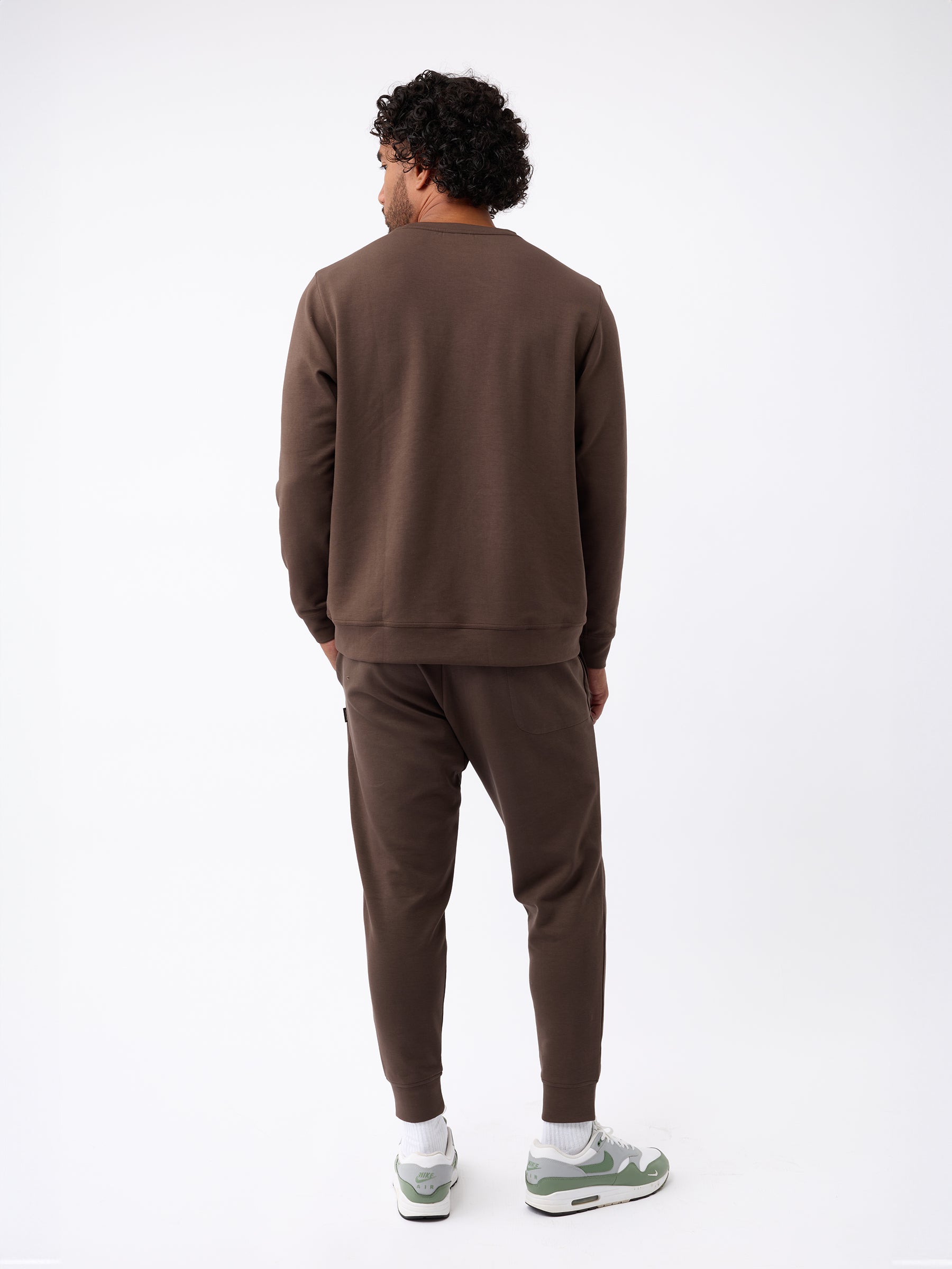 A man with curly hair stands facing away from the camera, donning a matching Men's StretchTech Crewneck and sweatpants in brown by Cozy Earth. He completes his outfit with white and green sneakers against a plain white background. |Color:Cacao