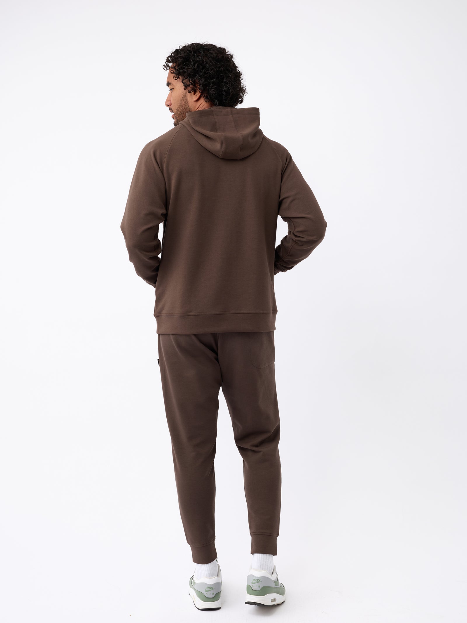 A man with curly hair is standing against a plain background, facing away from the camera. He is wearing Cozy Earth's Men's StretchTech Hoodie in brown, matching brown joggers, and light-colored sneakers. His hands are in the pockets and he appears to be looking slightly to his left. 