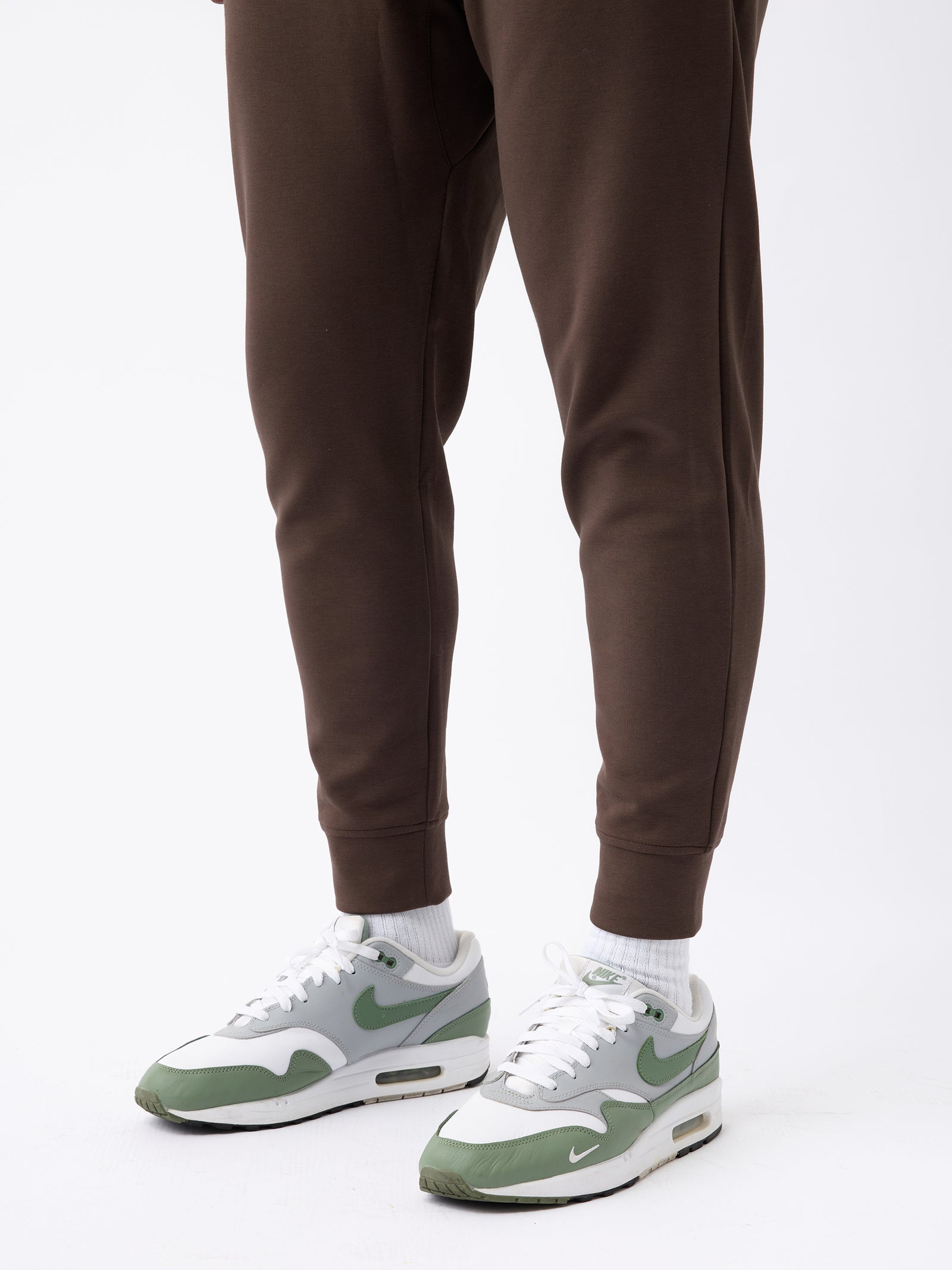 A person wearing Men's StretchTech Jogger in brown from Cozy Earth and white socks is shown from the knees down. They are also wearing a pair of gray and green Nike Air Max sneakers on a plain white background. 