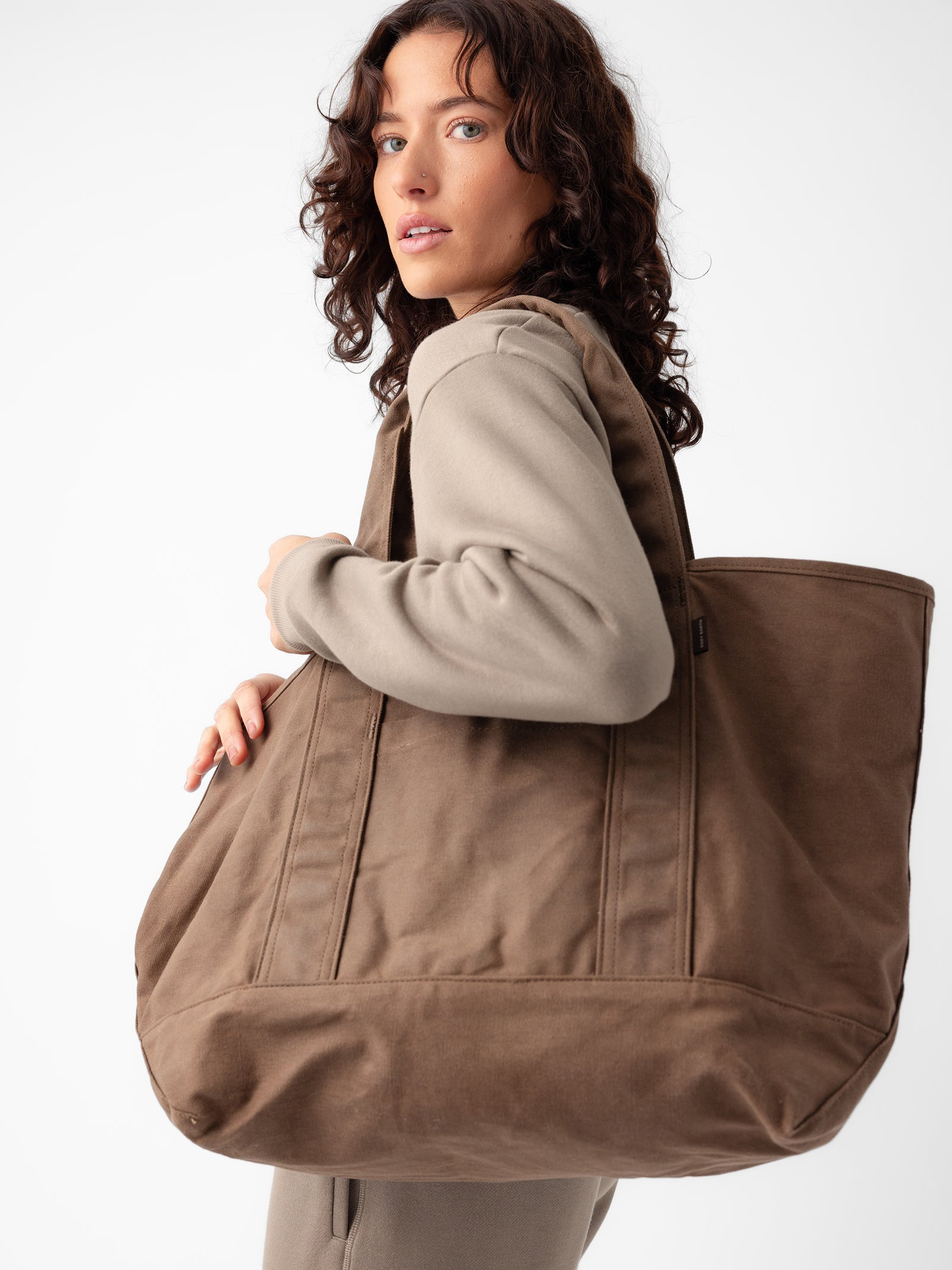 A person with shoulder-length wavy hair is looking over their shoulder while carrying a large Waxed Canvas Tote by Cozy Earth. The individual is wearing a beige outfit, and the background is plain white. 