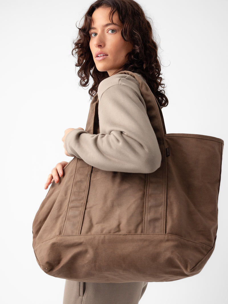 A person with shoulder-length wavy hair is looking over their shoulder while carrying a large Waxed Canvas Tote by Cozy Earth. The individual is wearing a beige outfit, and the background is plain white.
