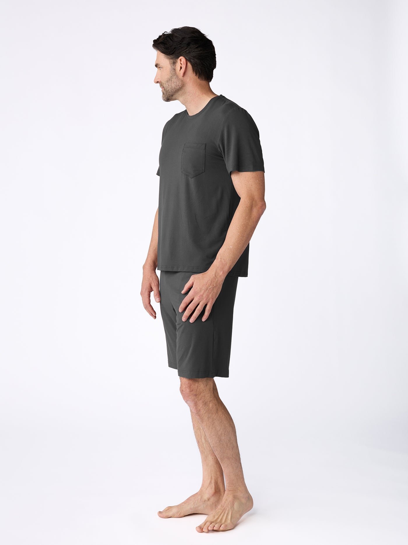 A man stands barefoot on a white background, facing left. He is wearing the Cozy Earth Men's Bamboo Stretch-Knit Short Sleeve Pajama Set—a matching dark gray shirt with a chest pocket and knee-length shorts. |Color:Charcoal