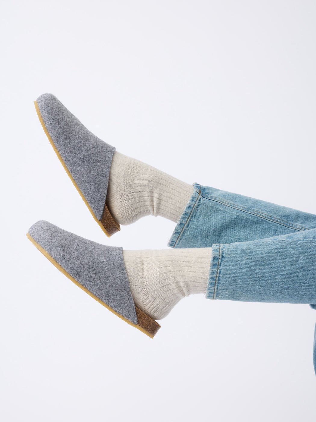 Someone wearing Cozy Earth's Lakehouse Clog gray felt slippers, beige ribbed socks, and light blue jeans crosses their legs slightly raised against a plain white background. 