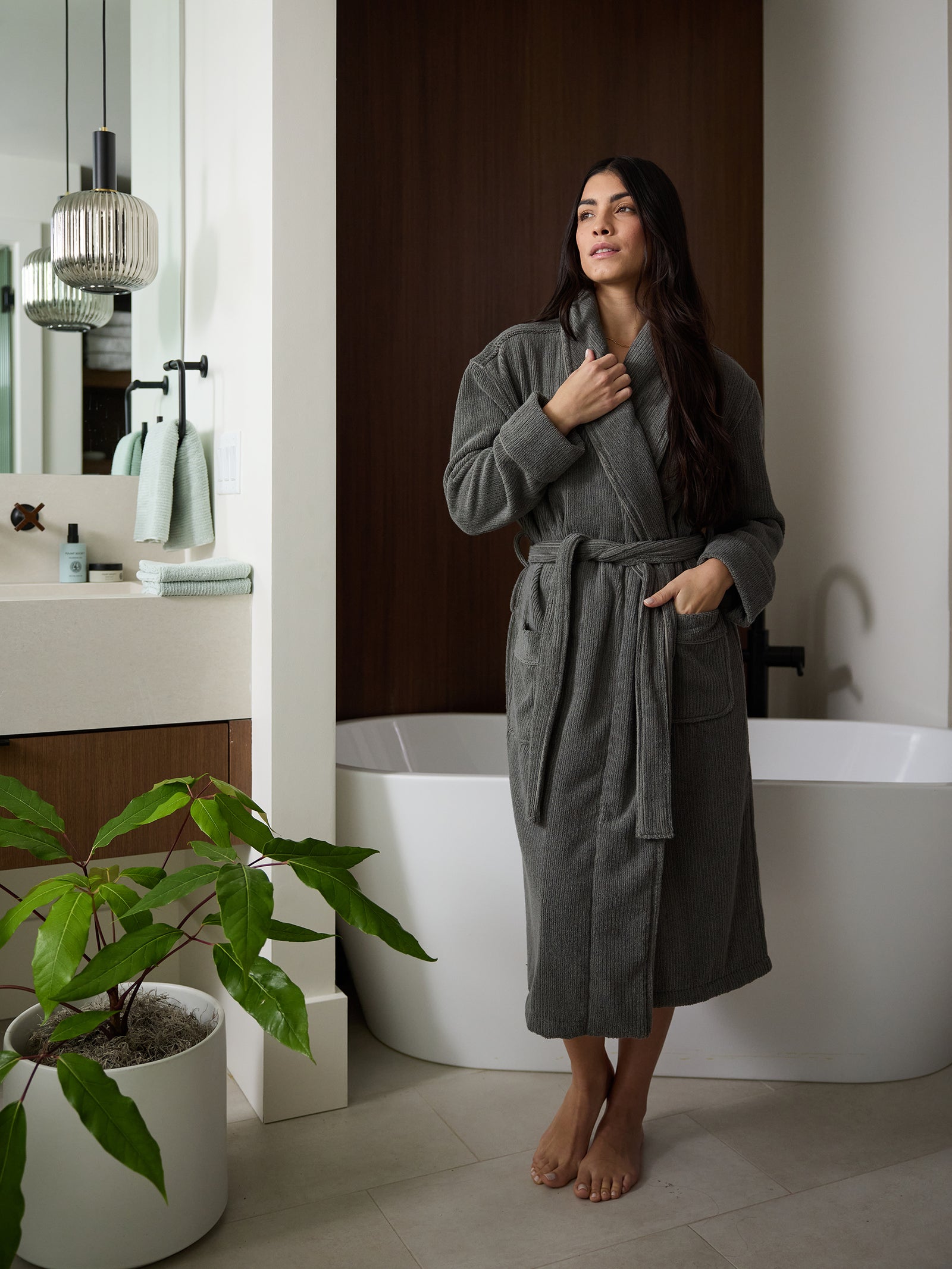 A barefoot woman wearing a Cozy Earth Ribbed Terry Bath Robe stands in a modern bathroom. She is holding her robe with one hand and looks thoughtfully into the distance. The bathroom features a white bathtub, a potted plant, towel on a rack, and a stylish pendant light. 