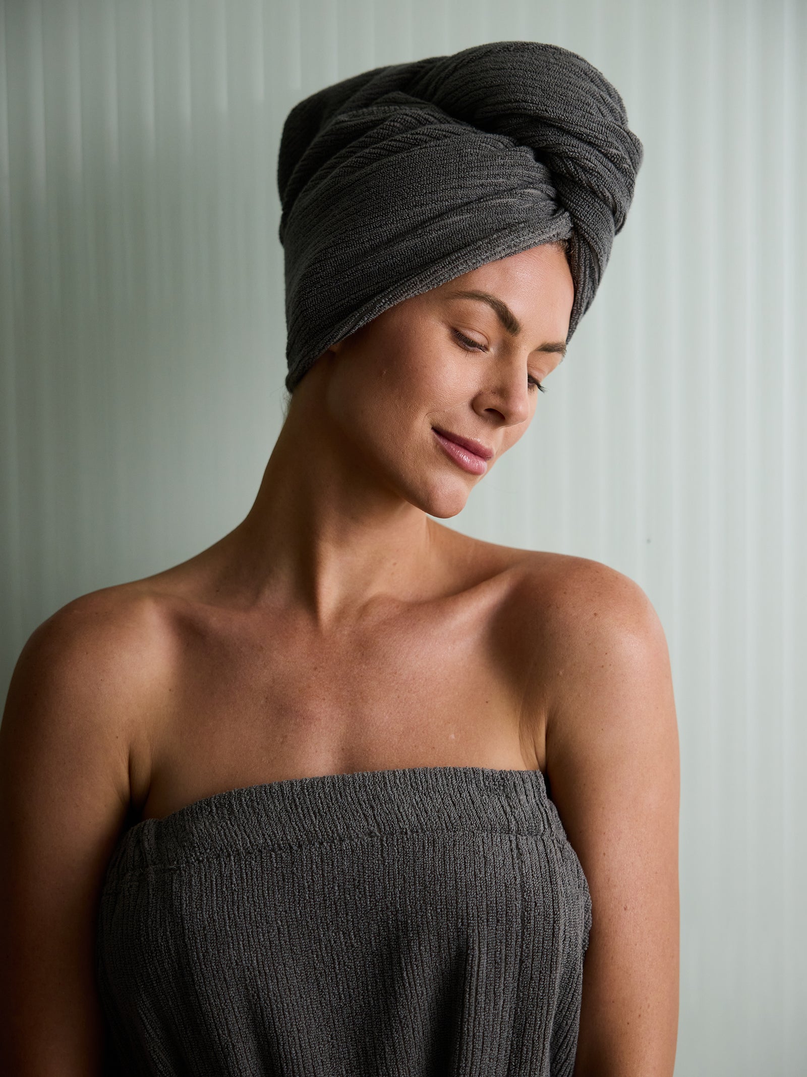 A person is wrapped in a dark towel with a Cozy Earth Ribbed Terry Hair Towel around their head, standing against a light-colored, vertically striped background. They have a relaxed expression, eyes closed and head tilted slightly to one side. 