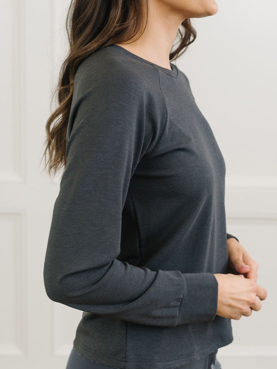 A person with long hair stands sideways in a dark gray Cozy Earth Women's Ultra-Soft Bamboo Wide Leg Pull On Pant & Pullover Crew Set, against a white paneled wall. 