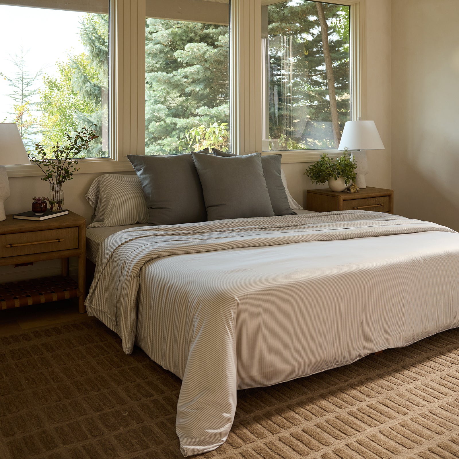 A cozy bedroom features a large bed adorned with a Bamboo Duvet Cover from Cozy Earth, white linens, and gray pillows. On either side, wooden nightstands hold lamps. Expansive windows offer views of lush green trees, while a textured rug adds warmth to the floor. 