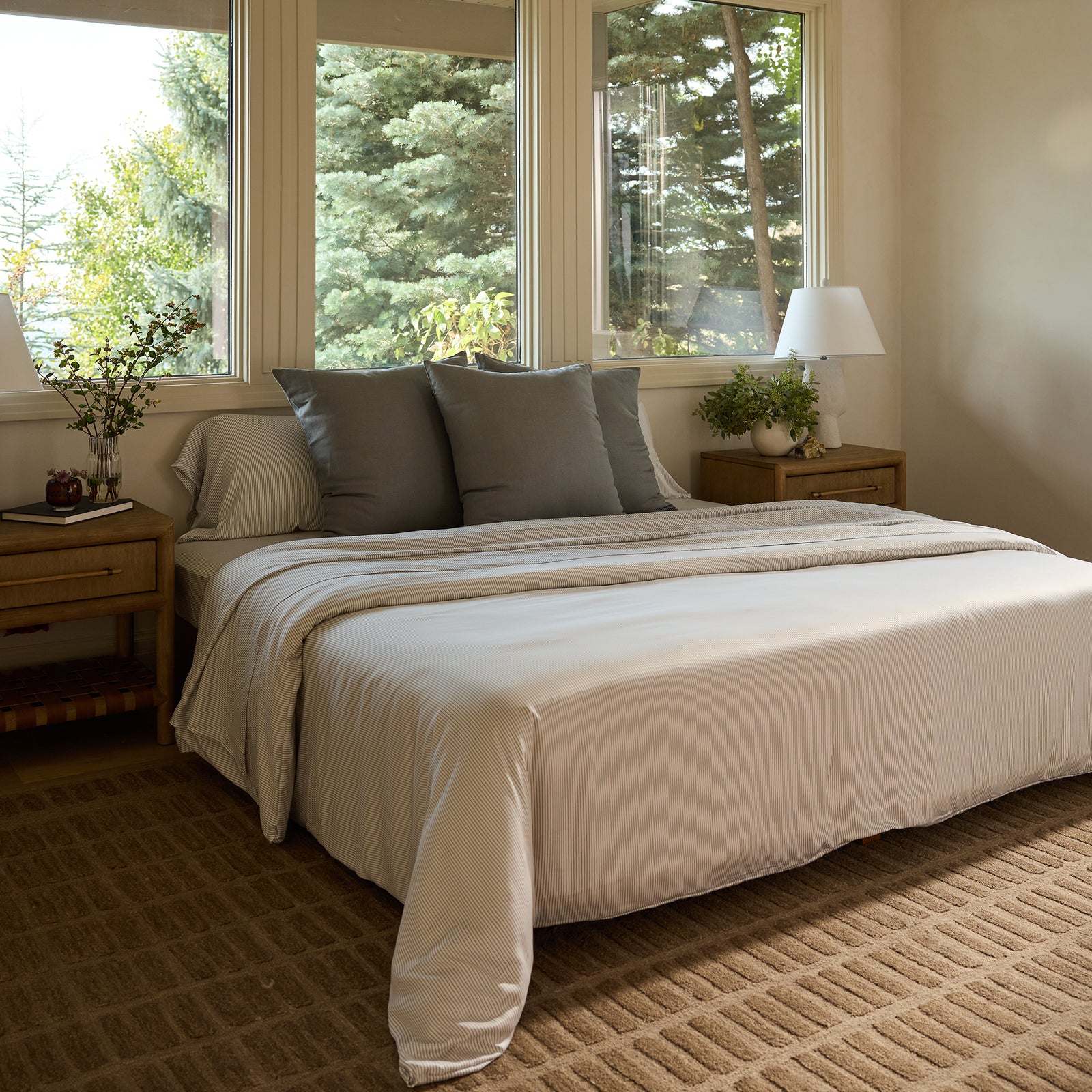 A cozy bedroom features a neatly made bed with Bamboo Fitted Sheets by Cozy Earth and gray pillows. Two wooden nightstands with lamps flank the bed. Large windows offer a view of lush greenery, and a textured rug covers the floor. 