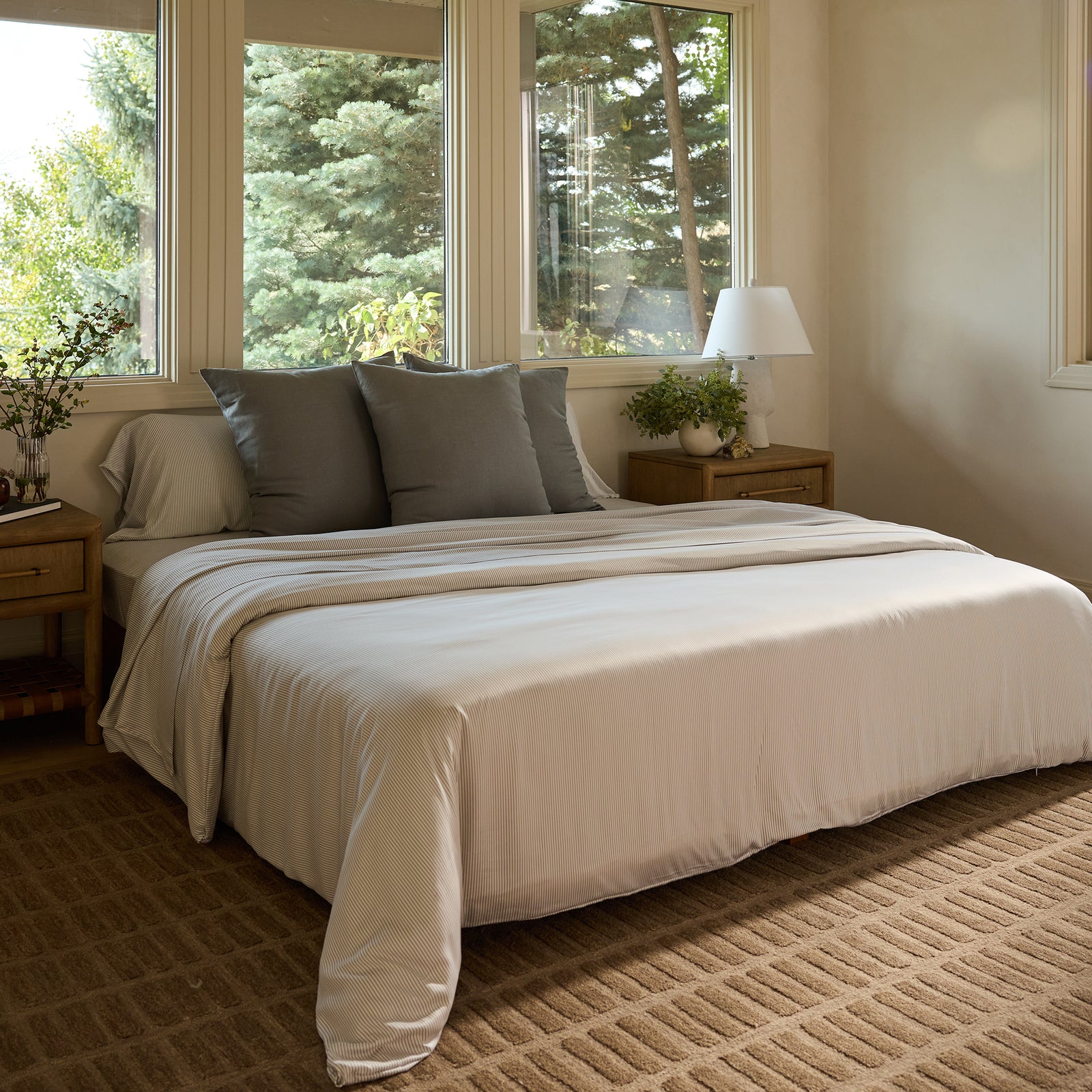 A cozy bedroom showcases a neatly made bed adorned with Cozy Earth's bamboo flat sheet in white, complemented by gray pillows. The room boasts two wooden nightstands equipped with lamps and has large windows that offer a view of verdant trees outside. The floor features a textured beige carpet. 