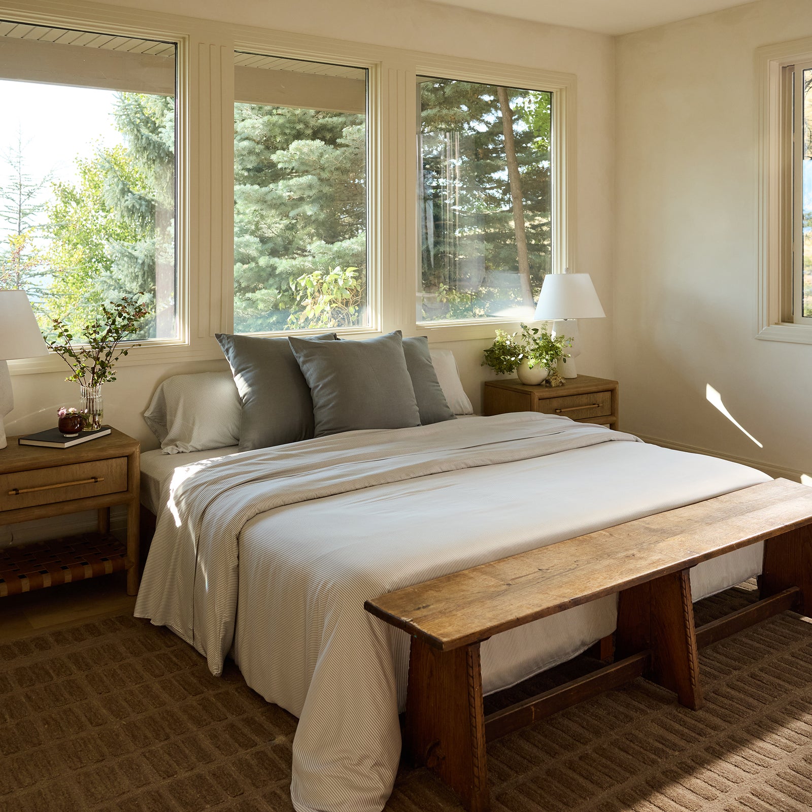 A comfortable bedroom features a spacious bed adorned with the Bamboo Sheet Set by Cozy Earth in white and gray tones. It's complemented by wooden nightstands holding white lamps and lush green plants. A wooden bench is positioned at the foot of the bed, while three large windows invite natural light from the trees outside. 