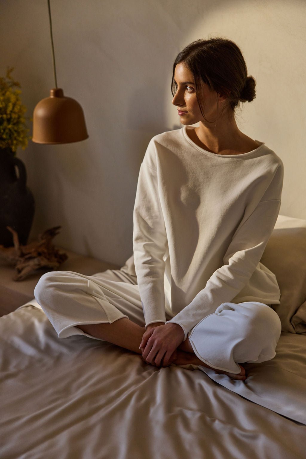 A person with dark hair tied back sits cross-legged on a bed, wearing a white long-sleeve shirt and Cozy Earth's Women's Chelsea Cropped Pant. They gaze thoughtfully to the side as warm light from a hanging lamp illuminates the room, highlighting a vase of yellow flowers in the background. 