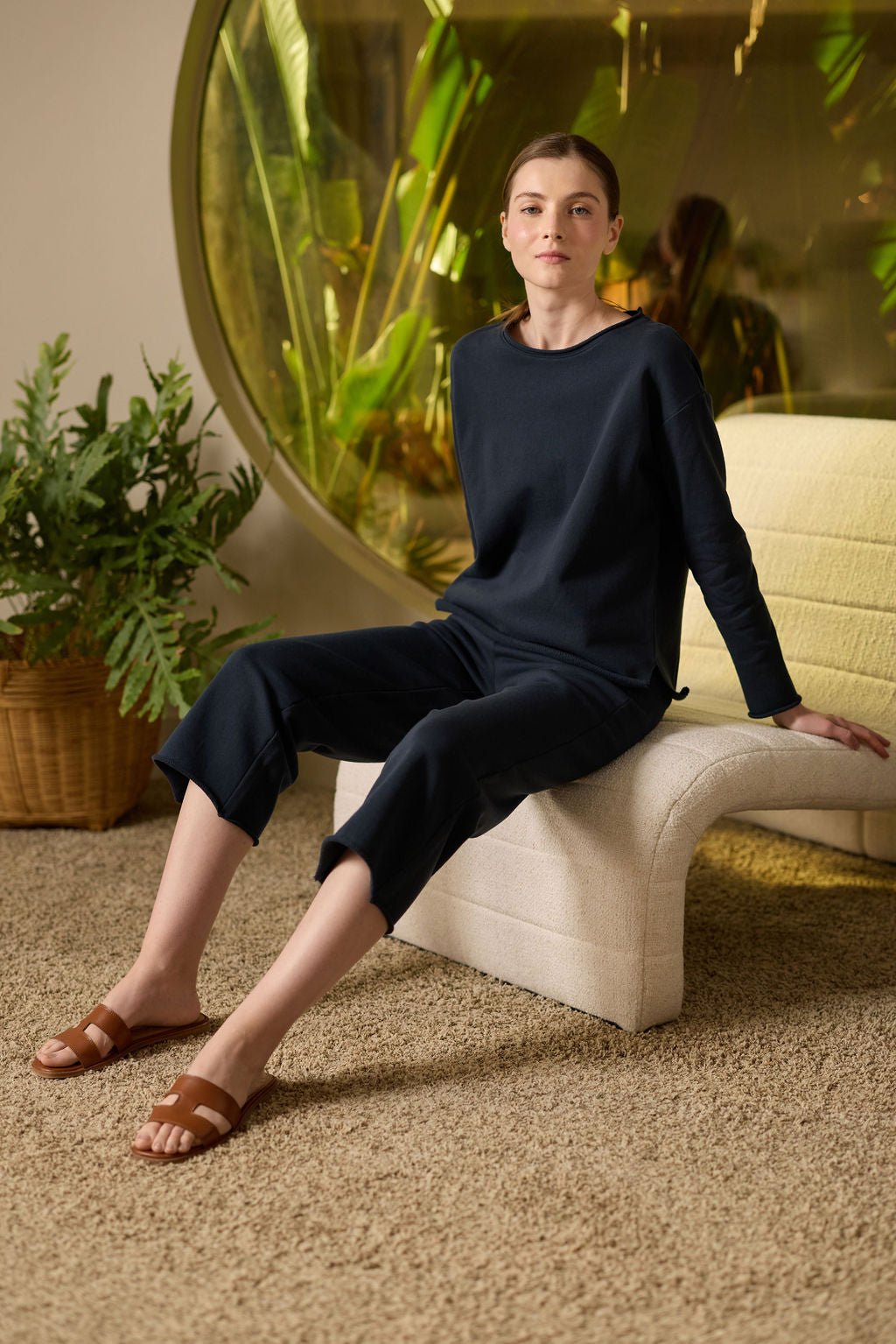 In a cozy room, someone sits on a modern white chair wearing a dark long-sleeve top, Cozy Earth's Women's Chelsea Cropped Pant, and brown sandals. A round window behind them reveals tropical plants, with a potted plant on the left and a soft beige carpet underfoot. 