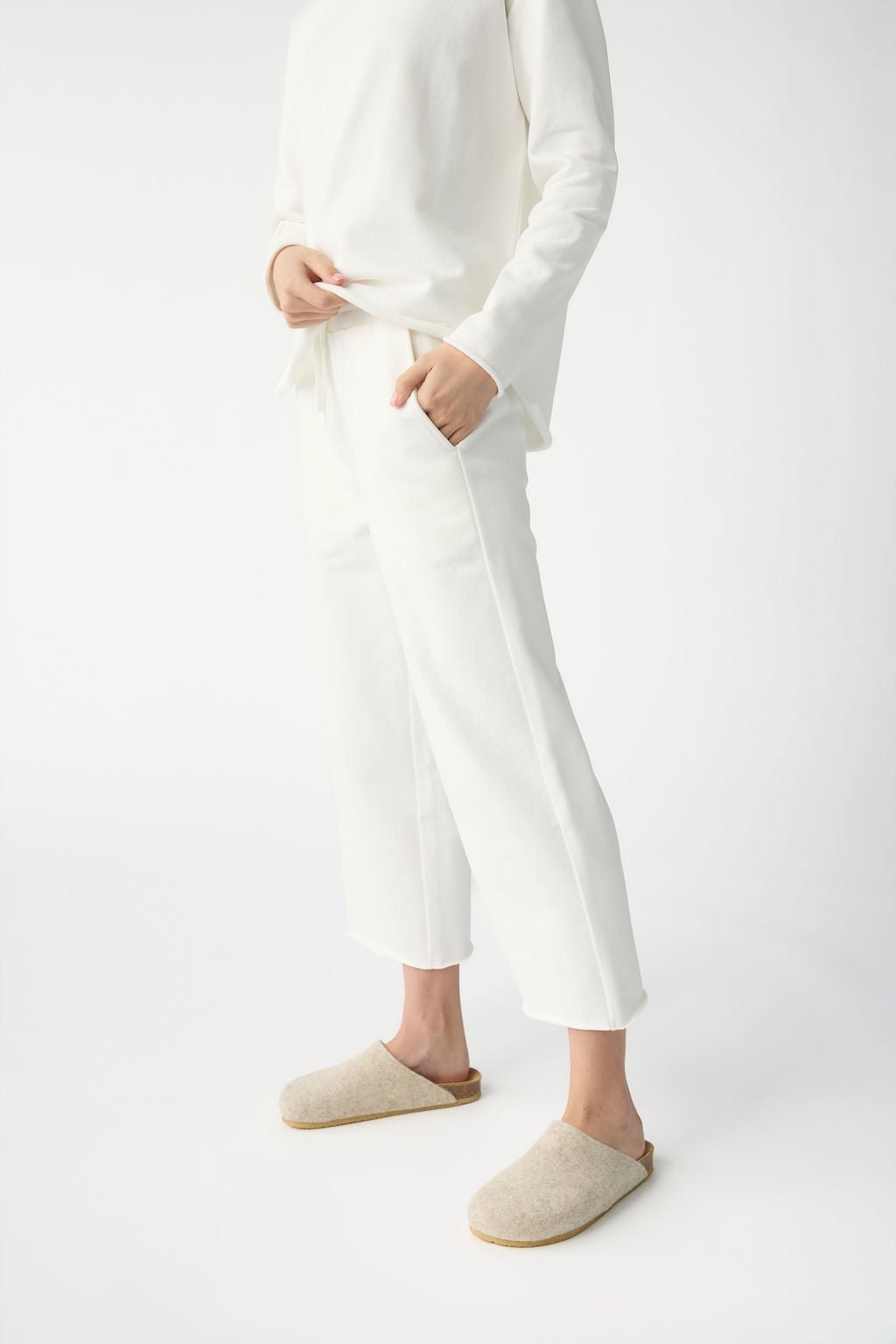 A person dressed in Cozy Earth's Women's Chelsea Set stands against a plain white background. The all-white outfit comprises a long-sleeve top and ankle-length pants, complemented by beige slippers, with hands tucked in pockets. 