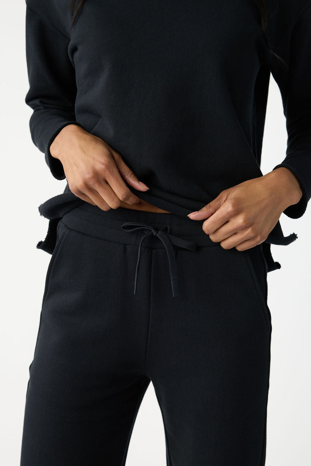 In a plain white setting, a person wearing Cozy Earth's Women's Chelsea Set in black adjusts the hem of the long-sleeve shirt to reveal their midriff. The matching joggers feature a drawstring waistband. 