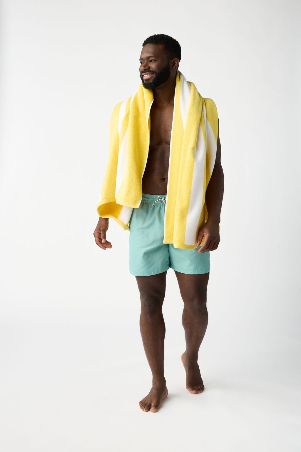 A man with a beard smiles, standing barefoot against a plain white background. He's wrapped in a Cozy Earth Classic Stripe Resort Towel and wearing light blue swim shorts. 