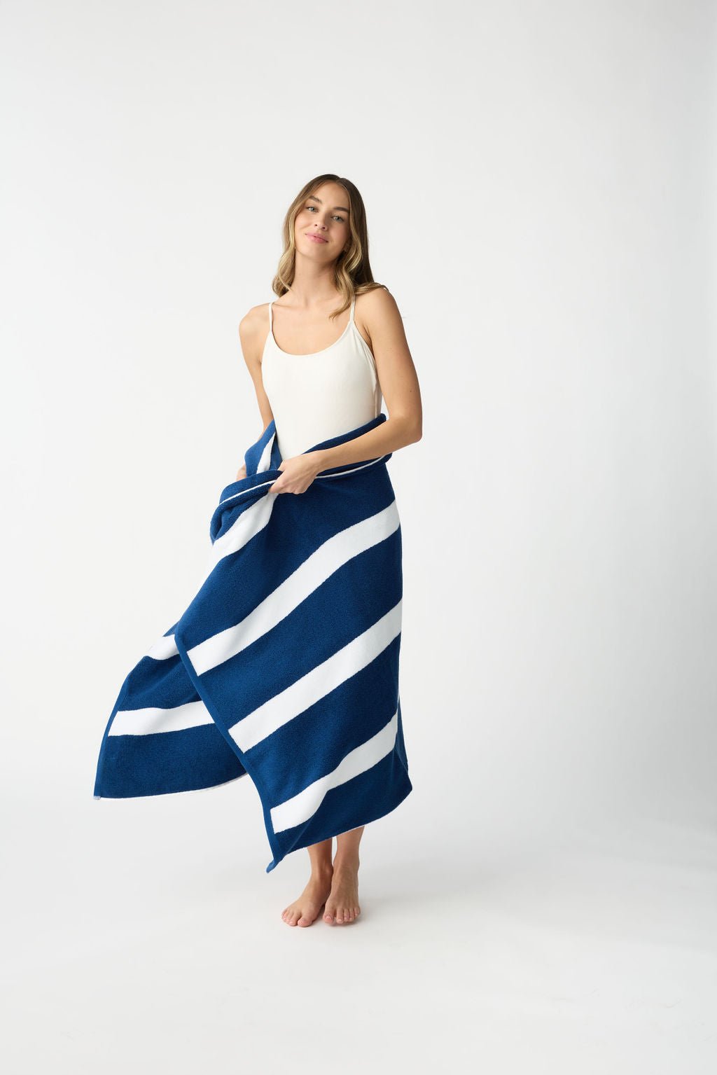 A woman stands barefoot on a white background, wrapped in the Cozy Earth Classic Stripe Resort Towel, draped gracefully around her sleeveless white outfit. Her hair is down, and she exudes a relaxed expression. |Color:Pacific