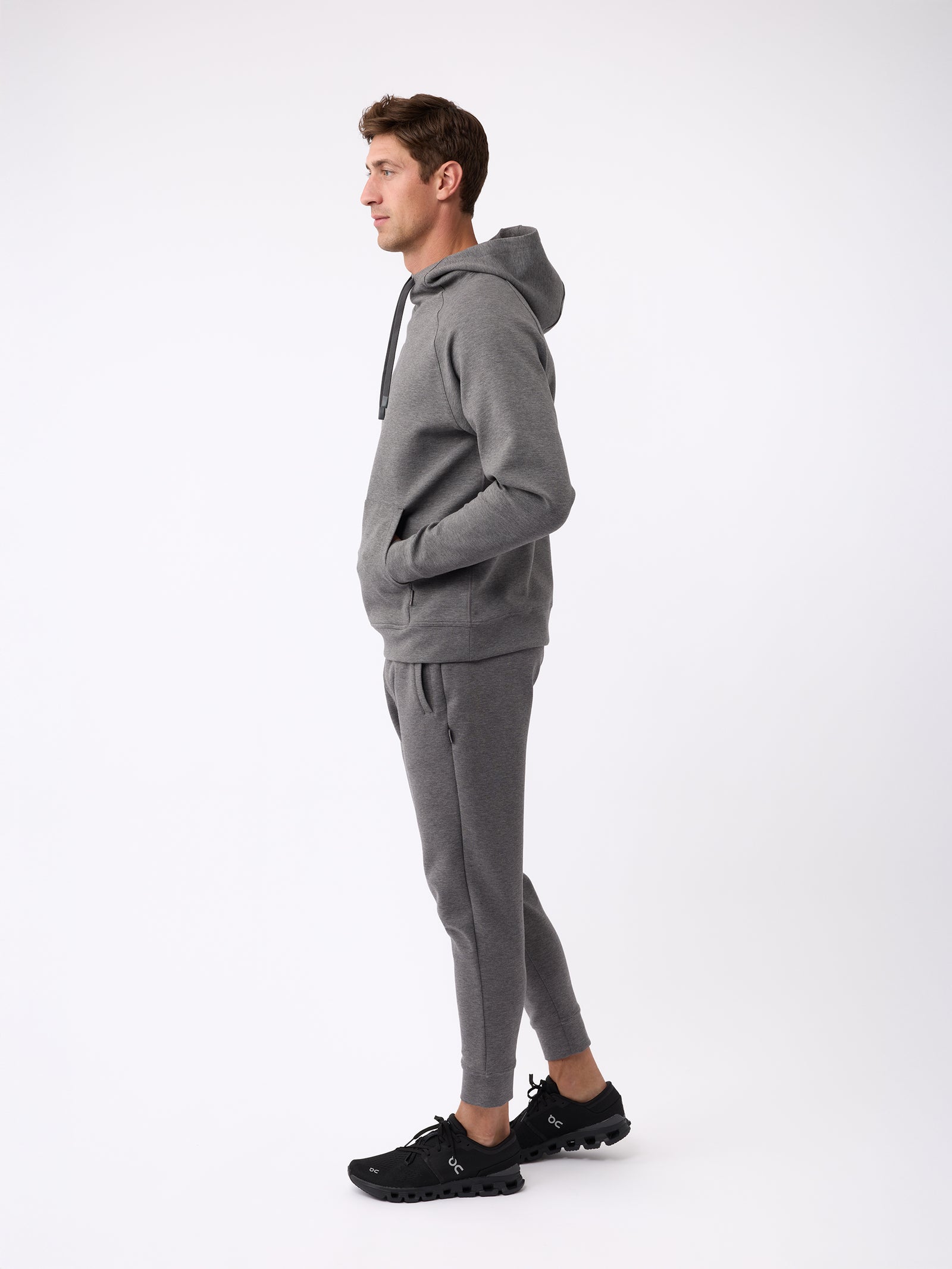 A person stands in profile against a white background, wearing the Cozy Earth Men's StretchTech Hoodie and matching joggers with black sneakers. They have their hands in the hoodie pockets and are looking forward. 