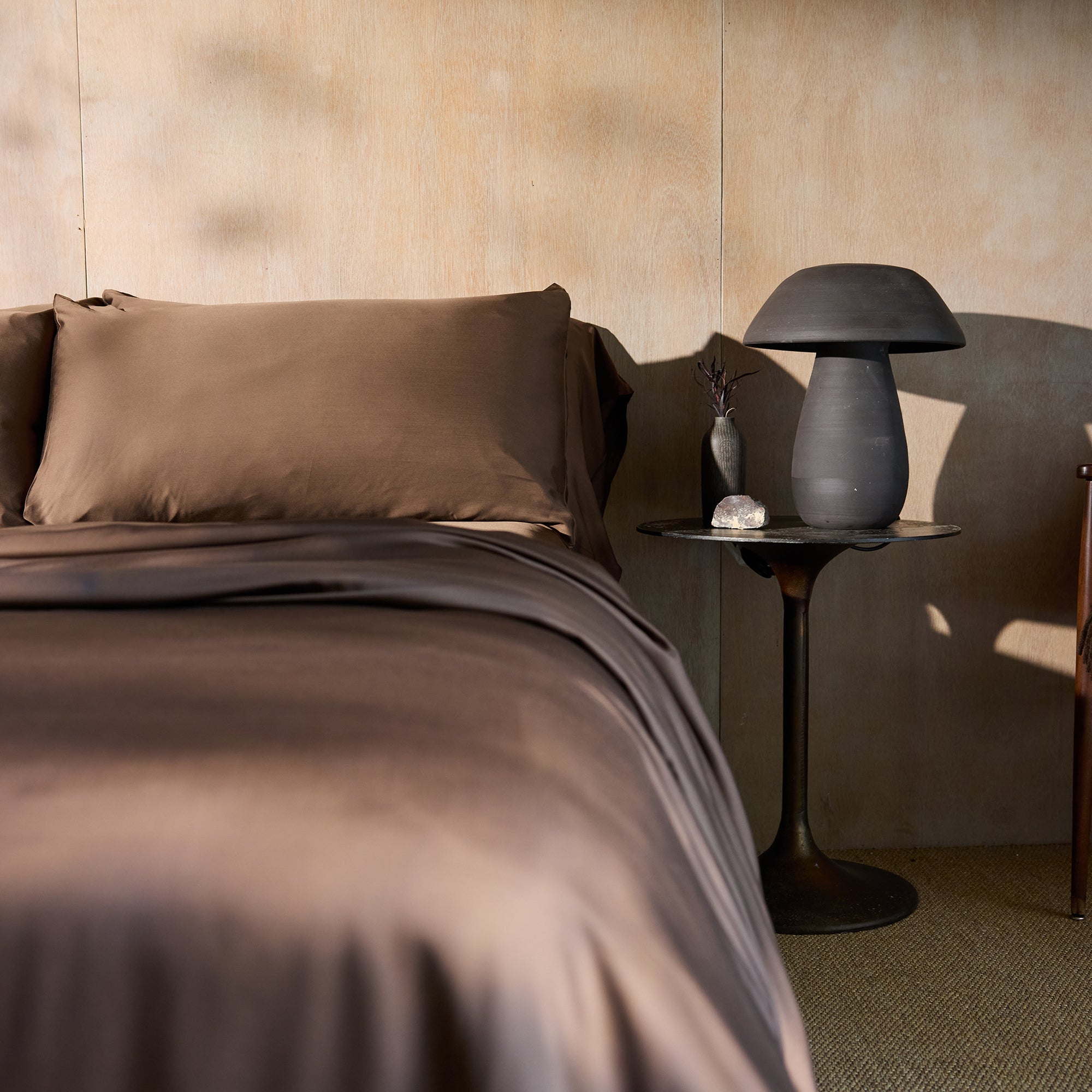 A cozy bedroom scene featuring a neatly made bed with Cozy Earth's Bamboo Sheet Set in brown, adorned with matching pillows. Next to the bed stands a round, black side table holding a modern, gray mushroom-shaped lamp, a small vase filled with dried flowers, and a decorative stone. The backdrop is defined by a wooden wall. 