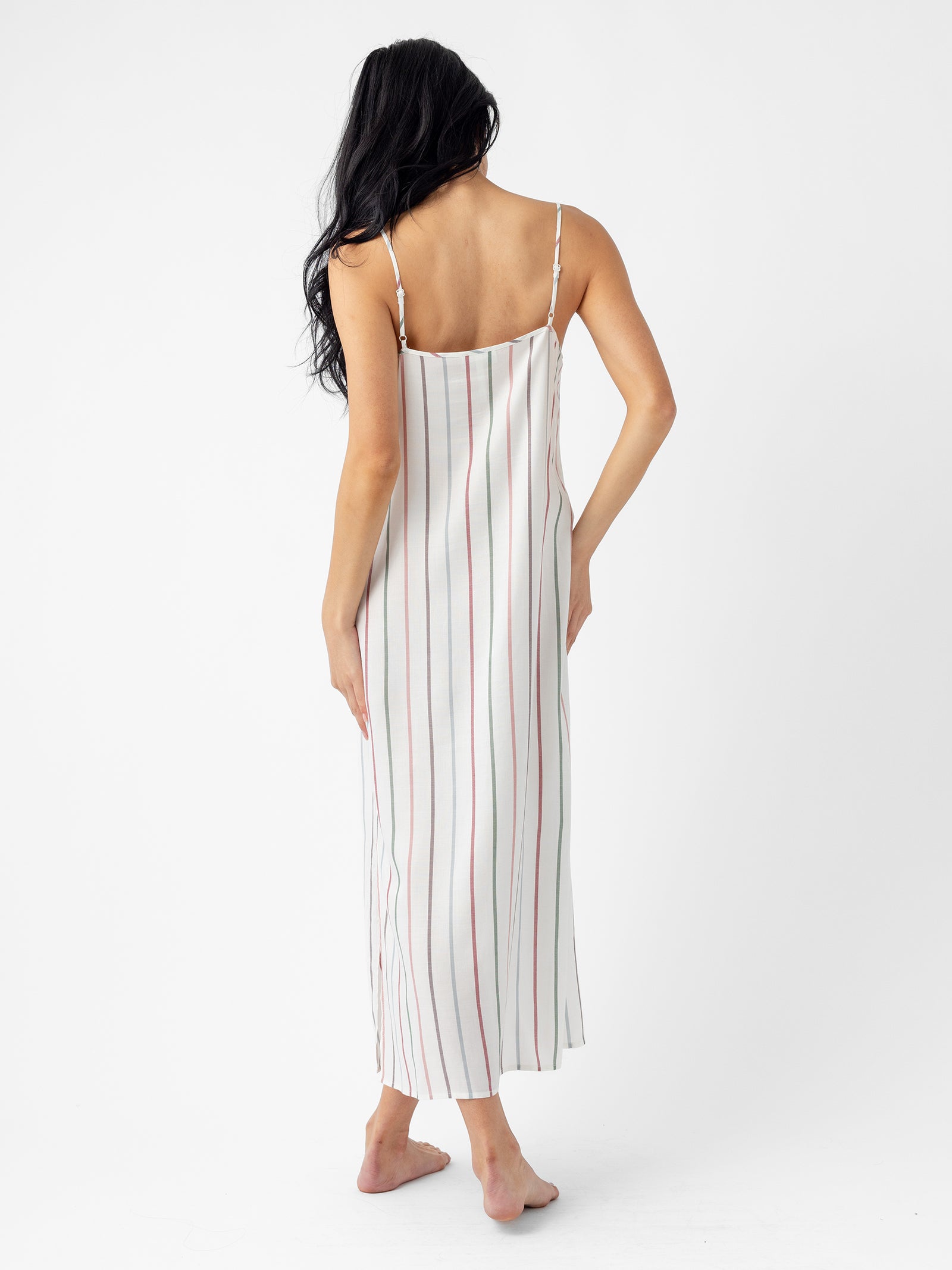 A person with long hair stands facing away, wearing the Women's Soft Woven Nightgown by Cozy Earth. The ankle-length garment features vertical stripes in red and black on a white background, has thin straps, and the individual is barefoot against a plain white backdrop. 