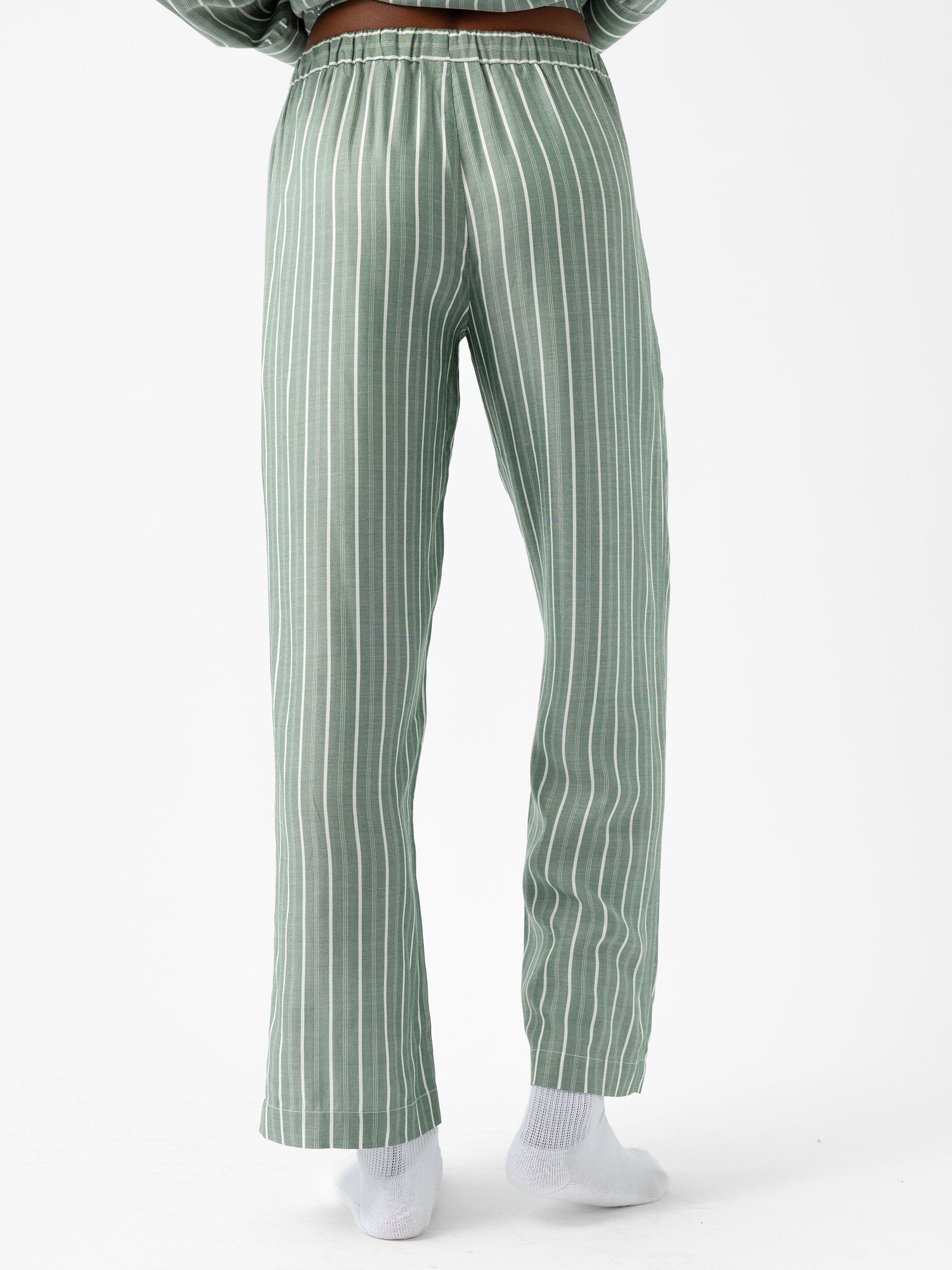 A person is standing against a plain white background, wearing Cozy Earth's Women's Soft Woven Pajama Pant in green and white vertical stripes, paired with white socks. Their upper body is not visible. 