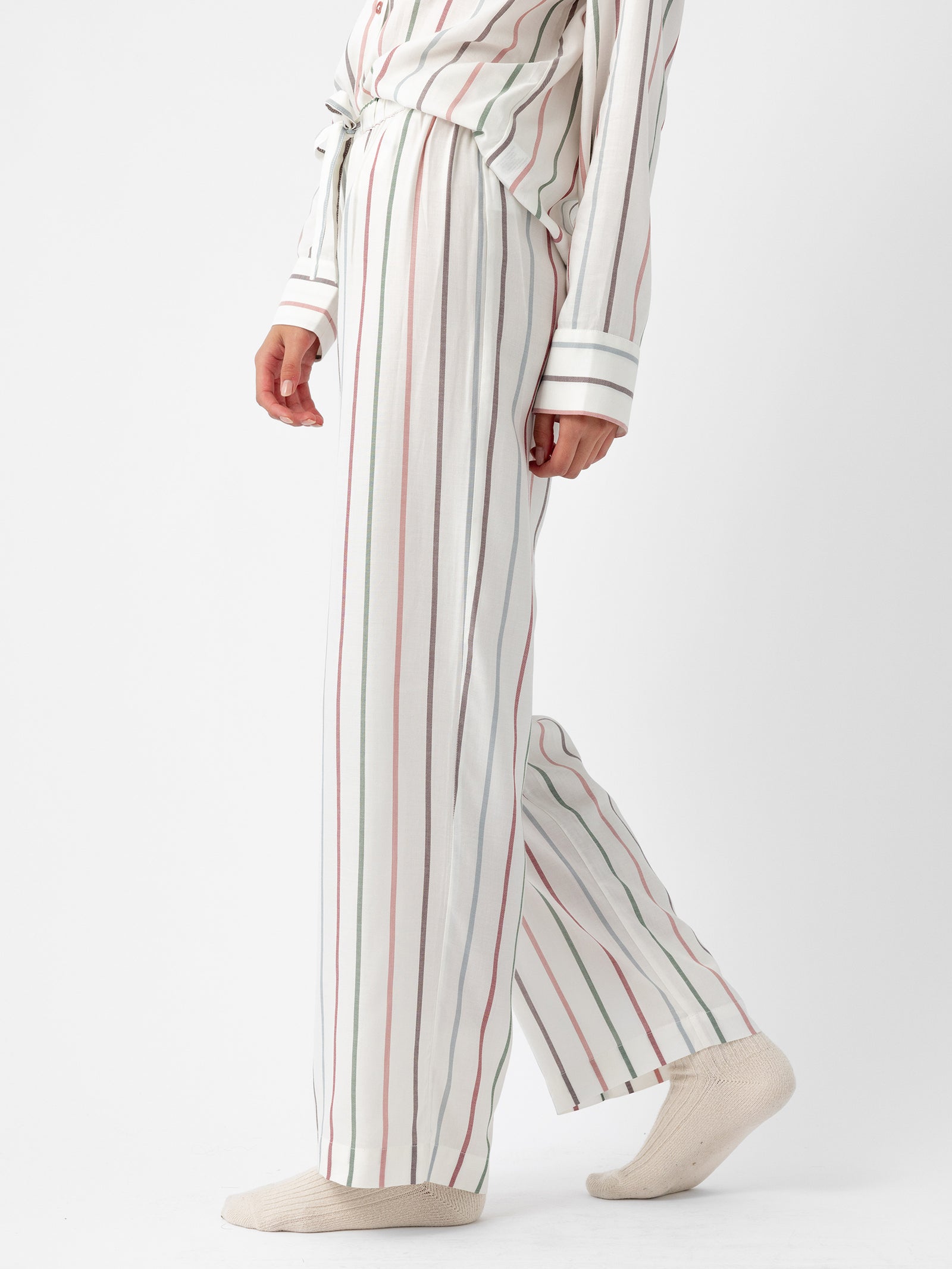 A person is wearing Cozy Earth's Women's Soft Woven Pajama Pant in a cream color with vertical stripes and a tied waist. The background is plain white, and the individual stands barefoot with one foot slightly ahead, showcasing a side view of the pajamas. 