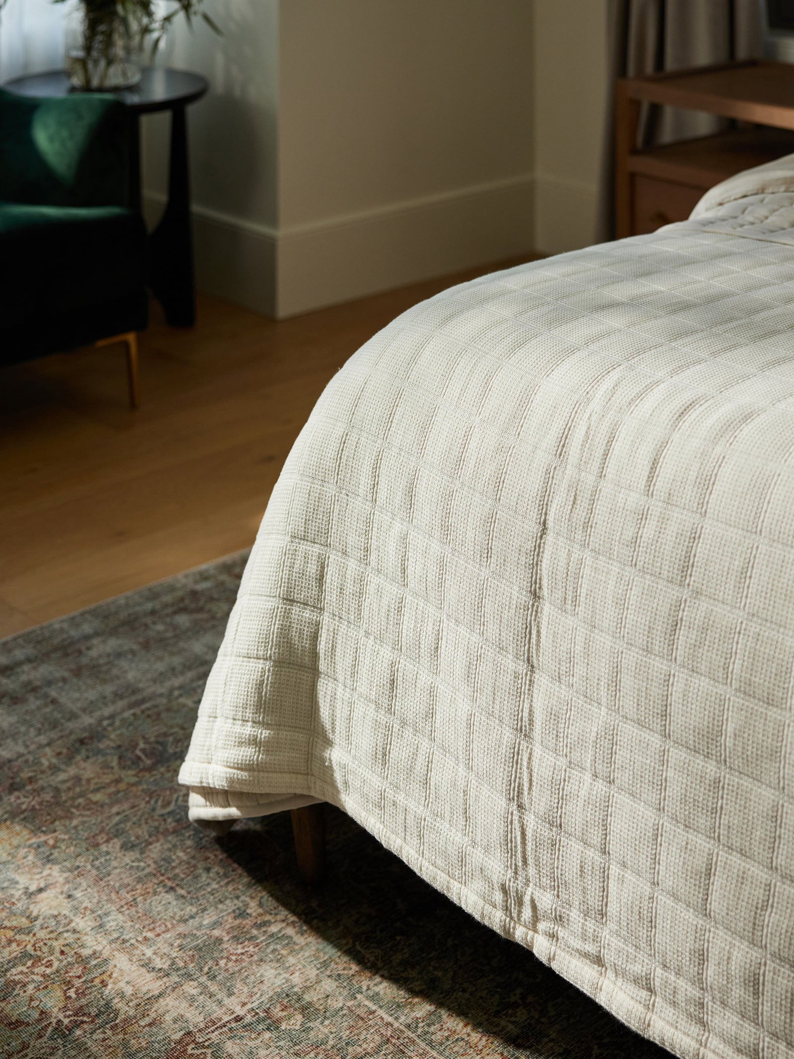 A cozy bedroom scene features a neatly made bed with a Cozy Earth Waffle Windowpane Blanket in off-white. Sunlight pours in, highlighting the textured blanket and part of a patterned rug. A dark green chair and a small side table are visible in the background. 