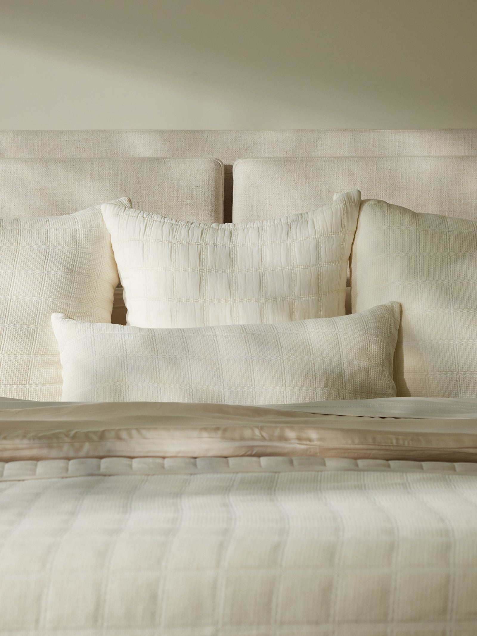 A neatly made bed with a beige textured headboard is perfectly complemented by soft, cream-colored linens. The bed features a quilted duvet and several pillows of varying sizes, including the exquisite Waffle Windowpane Pillow by Cozy Earth, all in coordinating creamy tones. The room is softly lit, creating a serene atmosphere. 