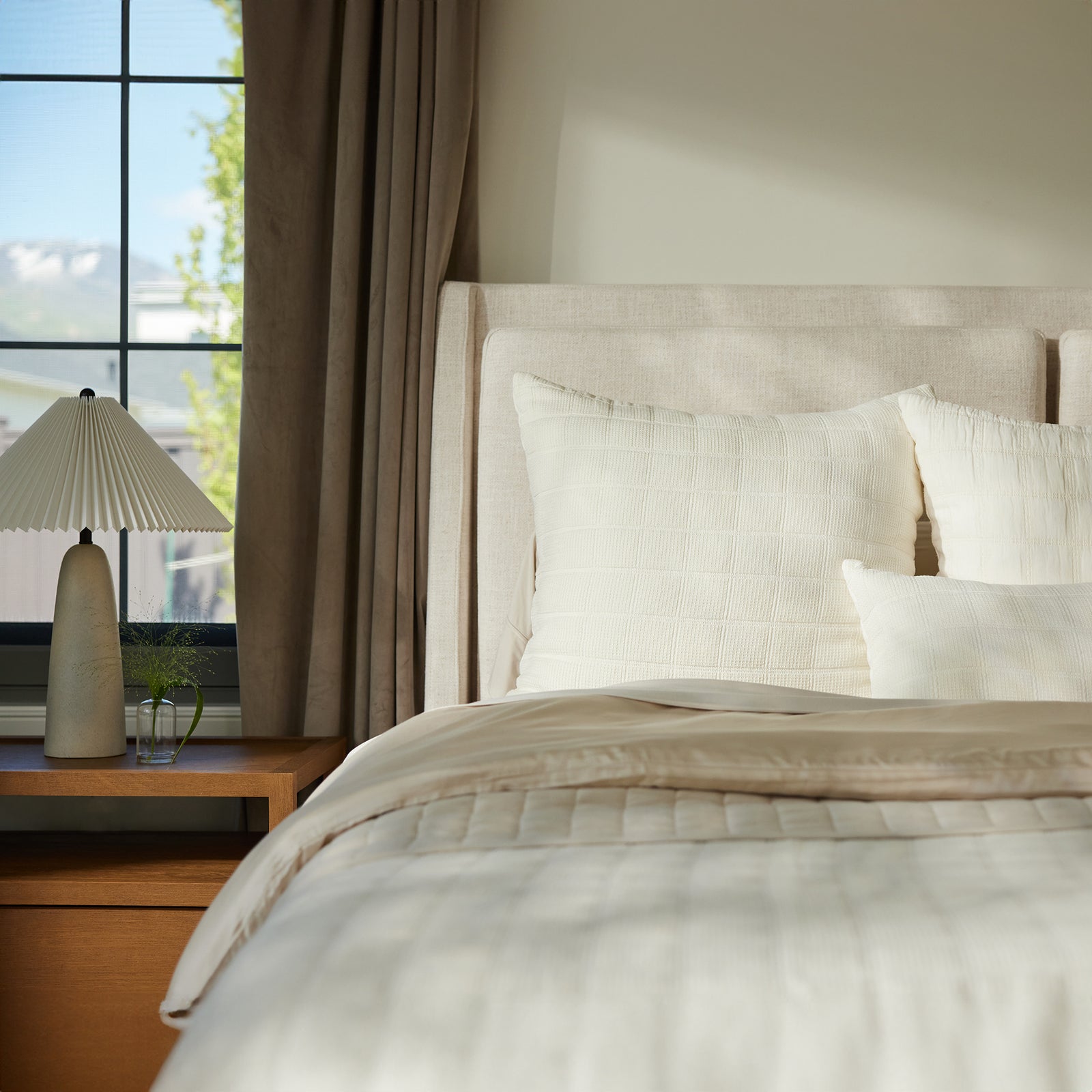 A neatly made bed adorned with the Cozy Earth Waffle Windowpane Pillow and beige and white bedding is situated next to a wooden nightstand, which is topped with a small decorative plant and a modern beige table lamp. A large window offering a view of the outdoors illuminates the scene. 