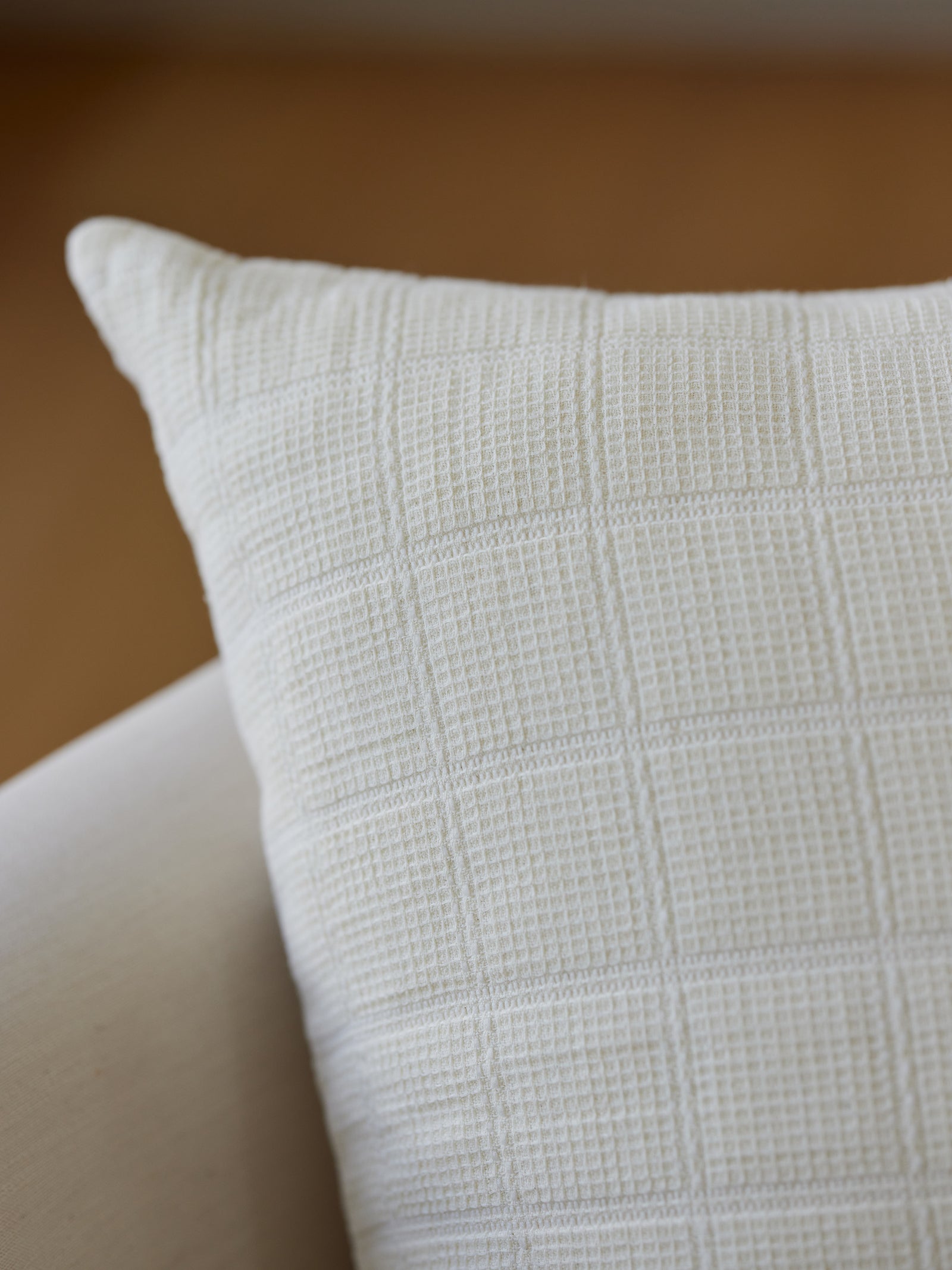 Close-up of the Cozy Earth Waffle Windowpane Pillow, featuring a textured beige fabric with a grid-like pattern. The pillow is placed on a light-colored sofa or chair against a blurred background, creating a cozy and minimalist ambiance. 