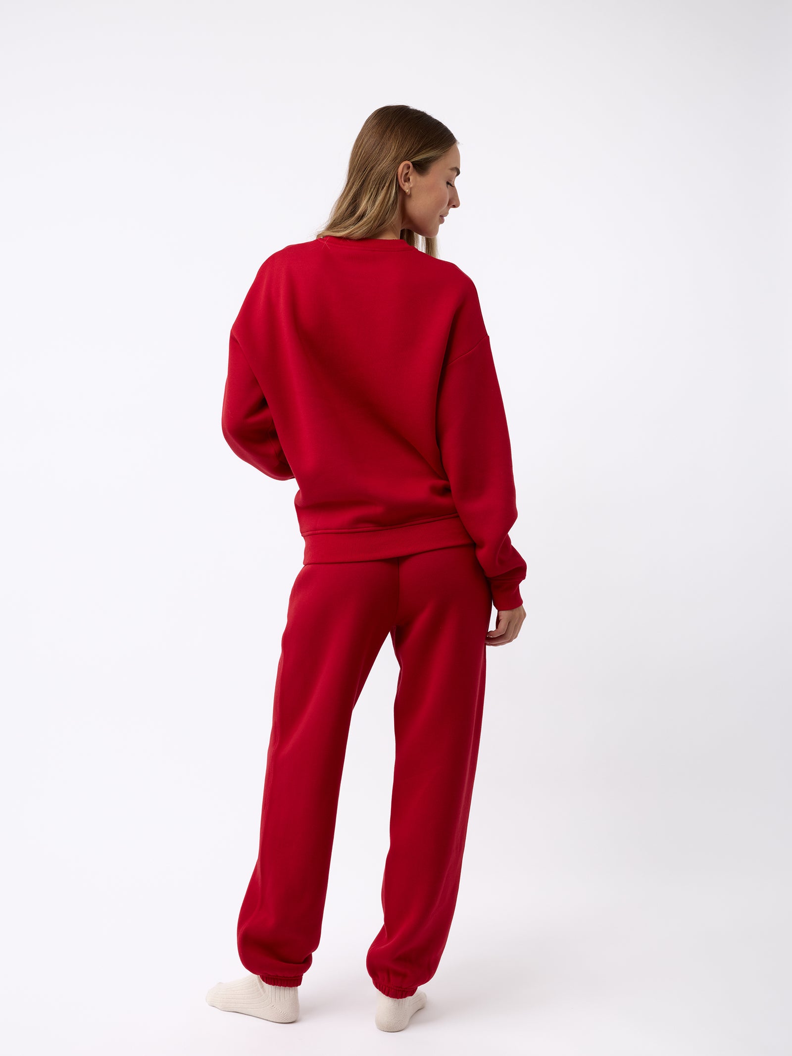 A person with long hair stands facing away, dressed in a red Women's CityScape Crewneck from Cozy Earth and beige socks, set against a plain white background. 