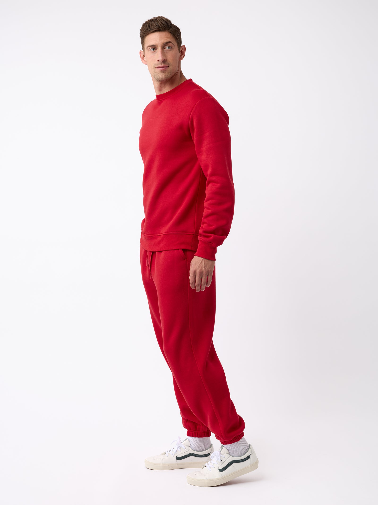 A man dressed in a red CityScape Crewneck by Cozy Earth stands against a white background, facing sideways. He is wearing white sneakers and looking slightly to the left. 