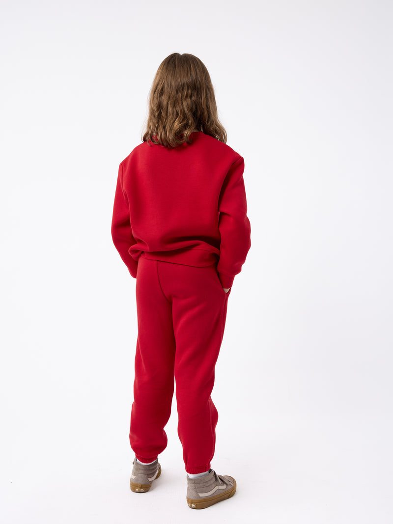 A child with medium-length brown hair is standing with their back to the camera, wearing a Kid's CityScape Crewneck by Cozy Earth in red and matching sweatpants, along with brown shoes. The background is plain white.