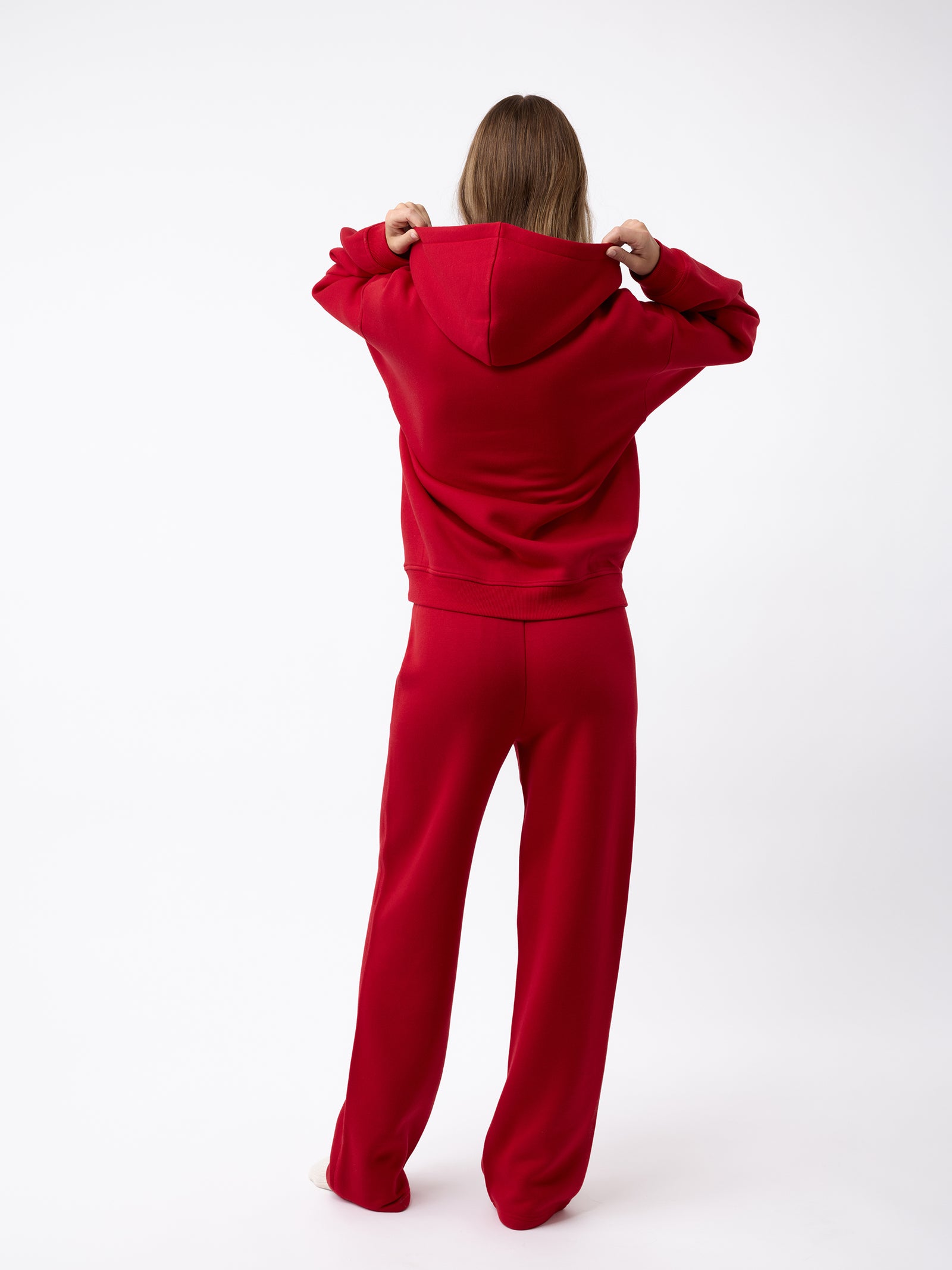 A person stands facing away, wearing a matching red Women's CityScape Hoodie and pants from Cozy Earth. They are holding the hood of the hoodie, set against a plain white background. 