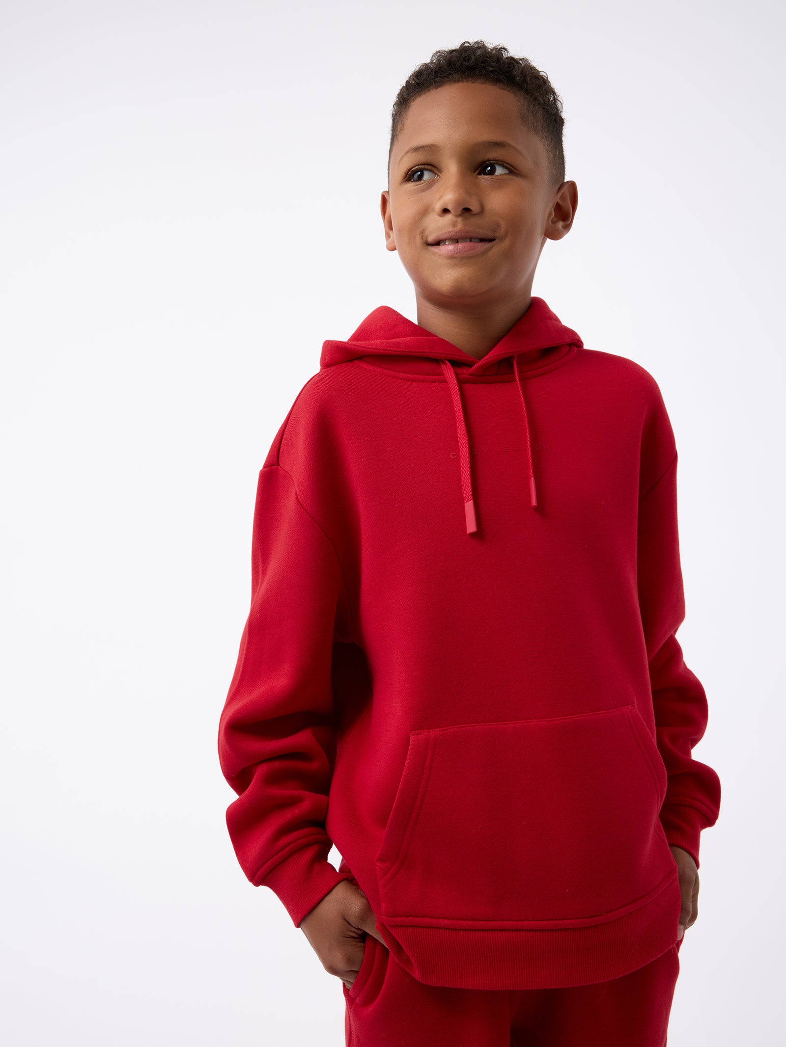 A young boy wearing the Kid's CityScape Hoodie from Cozy Earth stands with his hands in the spacious front pocket. He is looking slightly to the side and smiling against a plain white background. 