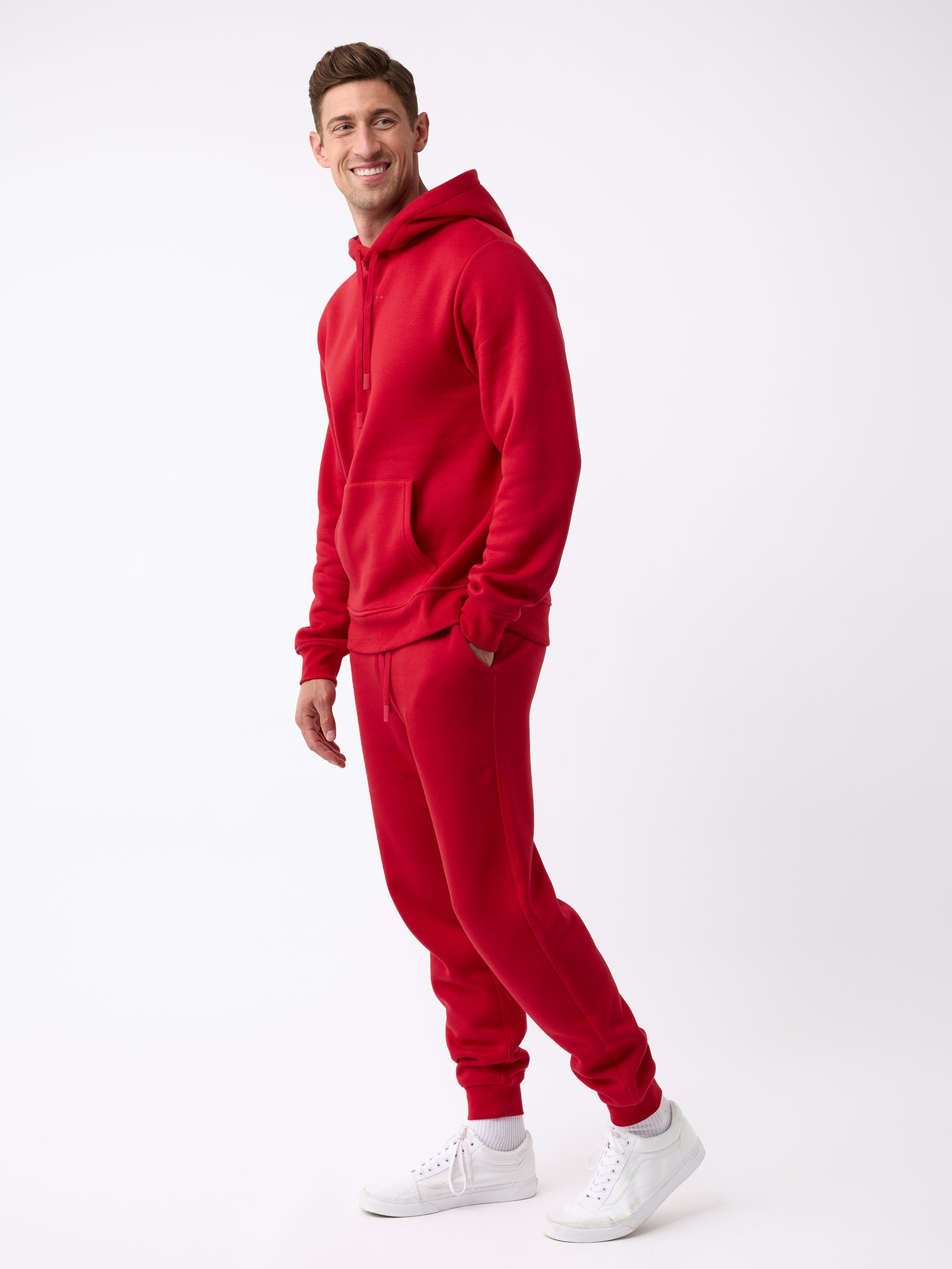 A person wearing a Men's CityScape Hoodie from Cozy Earth in a vibrant red color, along with coordinating sweatpants, stands against a plain white background. They are smiling with their hands in the hoodie pockets and wearing white sneakers. 