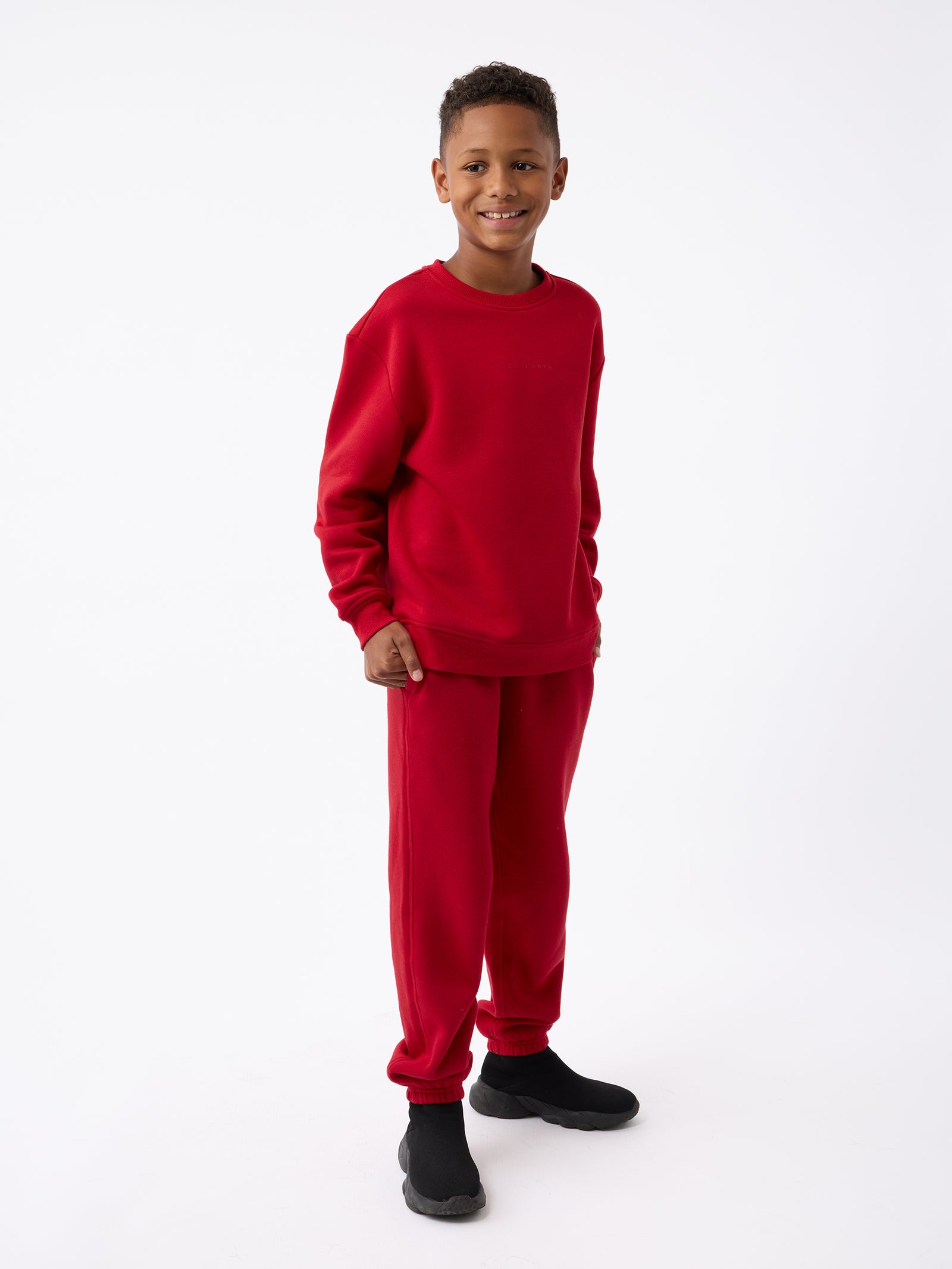 A young boy smiles while dressed in the Kid's CityScape Sweatpant, a matching red sweatshirt and sweatpants set from Cozy Earth, paired with black shoes. He stands against a plain white background. 