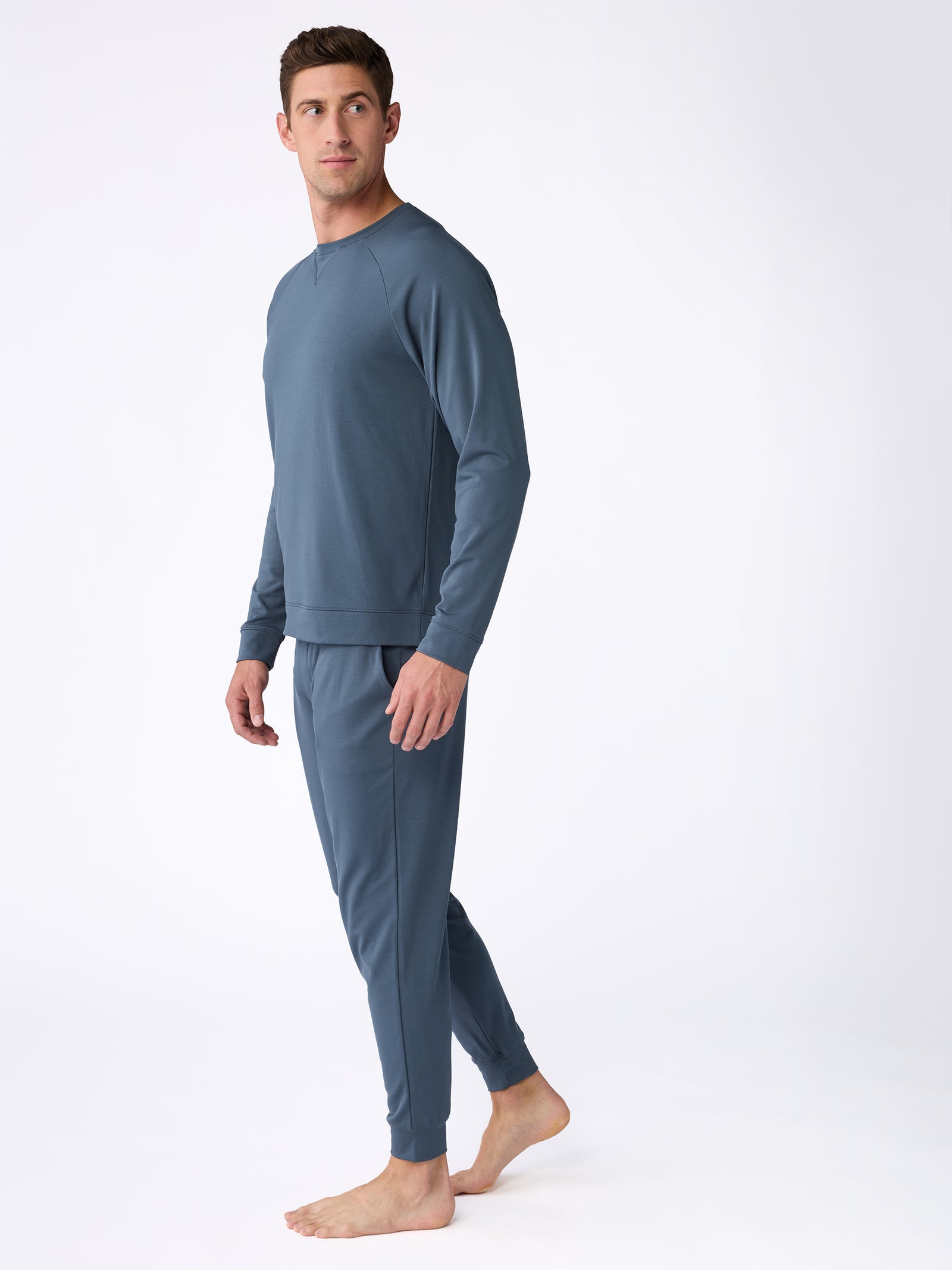 A person is barefoot on a white background, wearing the Men's Ultra-Soft Bamboo Pullover Crew by Cozy Earth in a matching blue set that includes long-sleeve tops and pants. The individual gazes to the side, one foot slightly forward, showcasing the outfit's comfortable and casual style. 